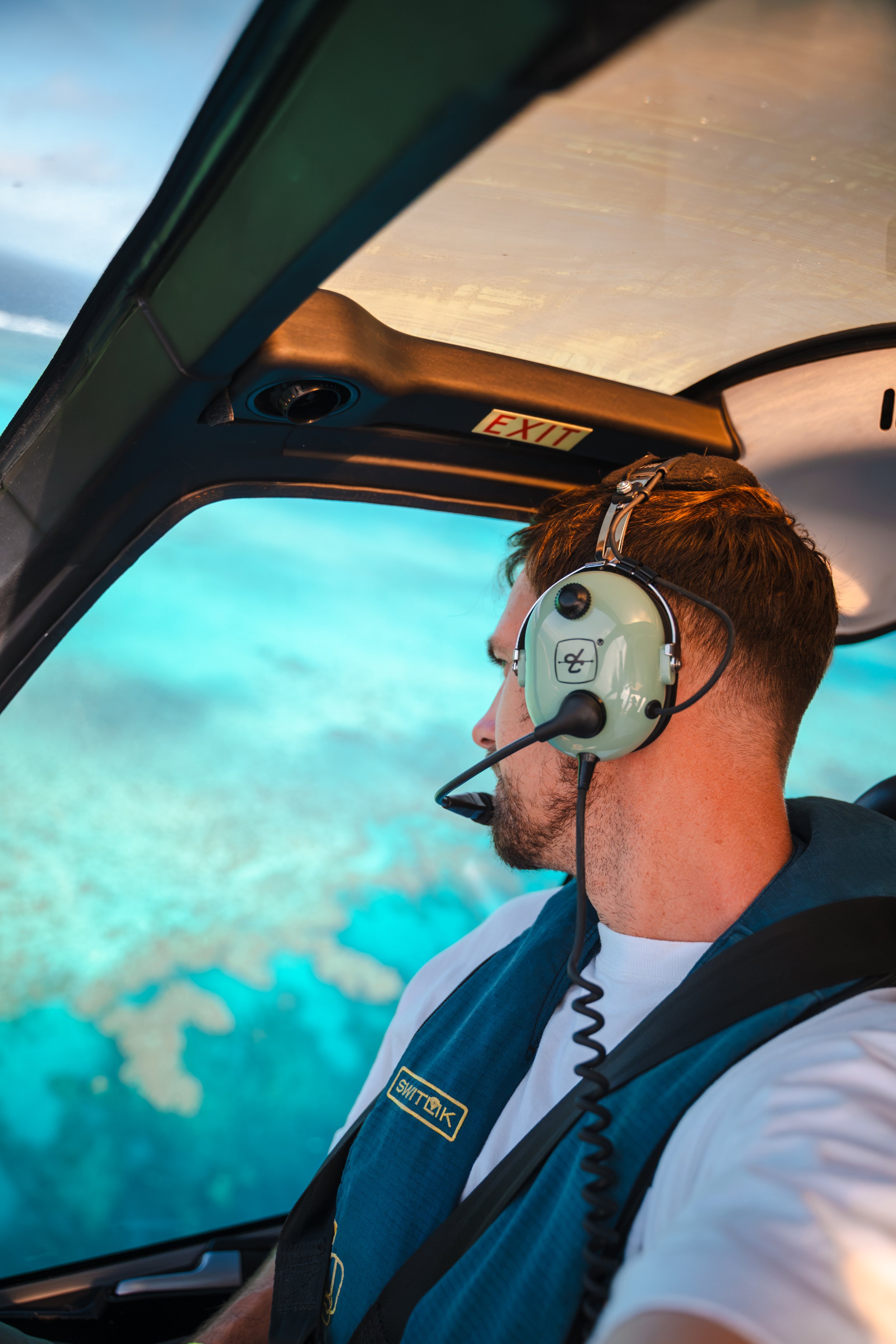 A male airline pilot is in the cockpit of an airplane, wearing a headset and a uniform. The background shows the cockpit window with a view of the ocean or sea.