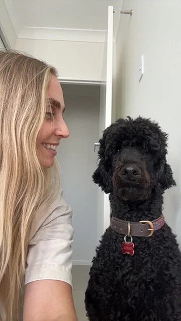 A woman smiling at a black curly-haired dog with a red collar, in a room with an open closet in the background.