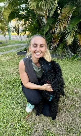 A woman crouching and smiling while hugging a black dog outdoors with large green tropical plants in the background.