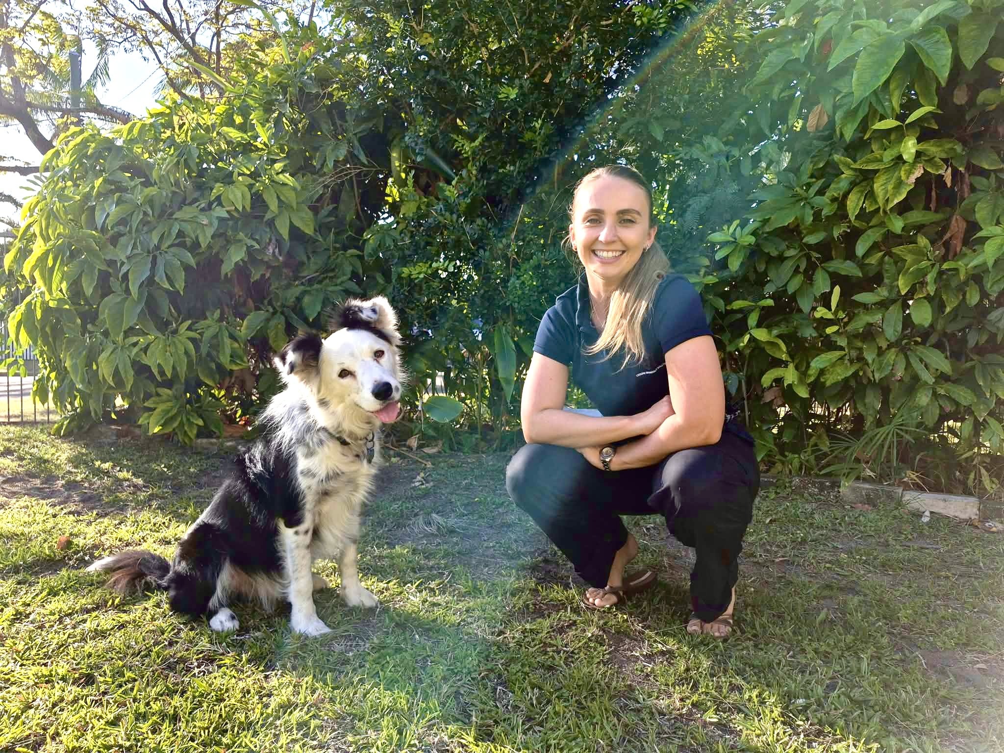 A woman with long blonde hair smiling and squatting next to a black and white Australian Shepherd dog with blue eyes in a lush green garden.