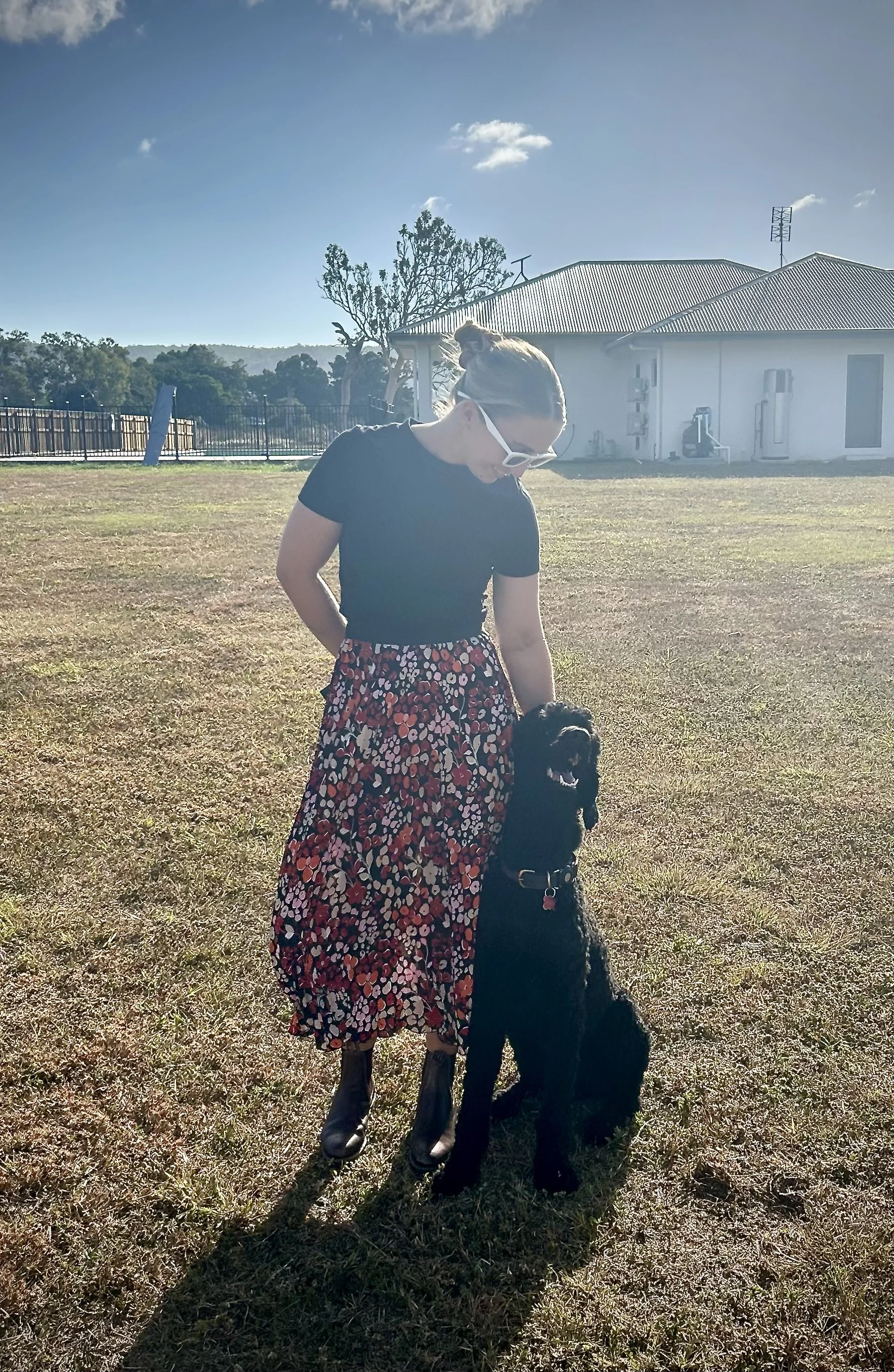 A woman with blonde hair tied up in a bun wearing white sunglasses and a black top, standing outdoors on a grassy area with a black dog, in front of a white building with a metal roof and a tree nearby. The sky is clear with a few clouds.
