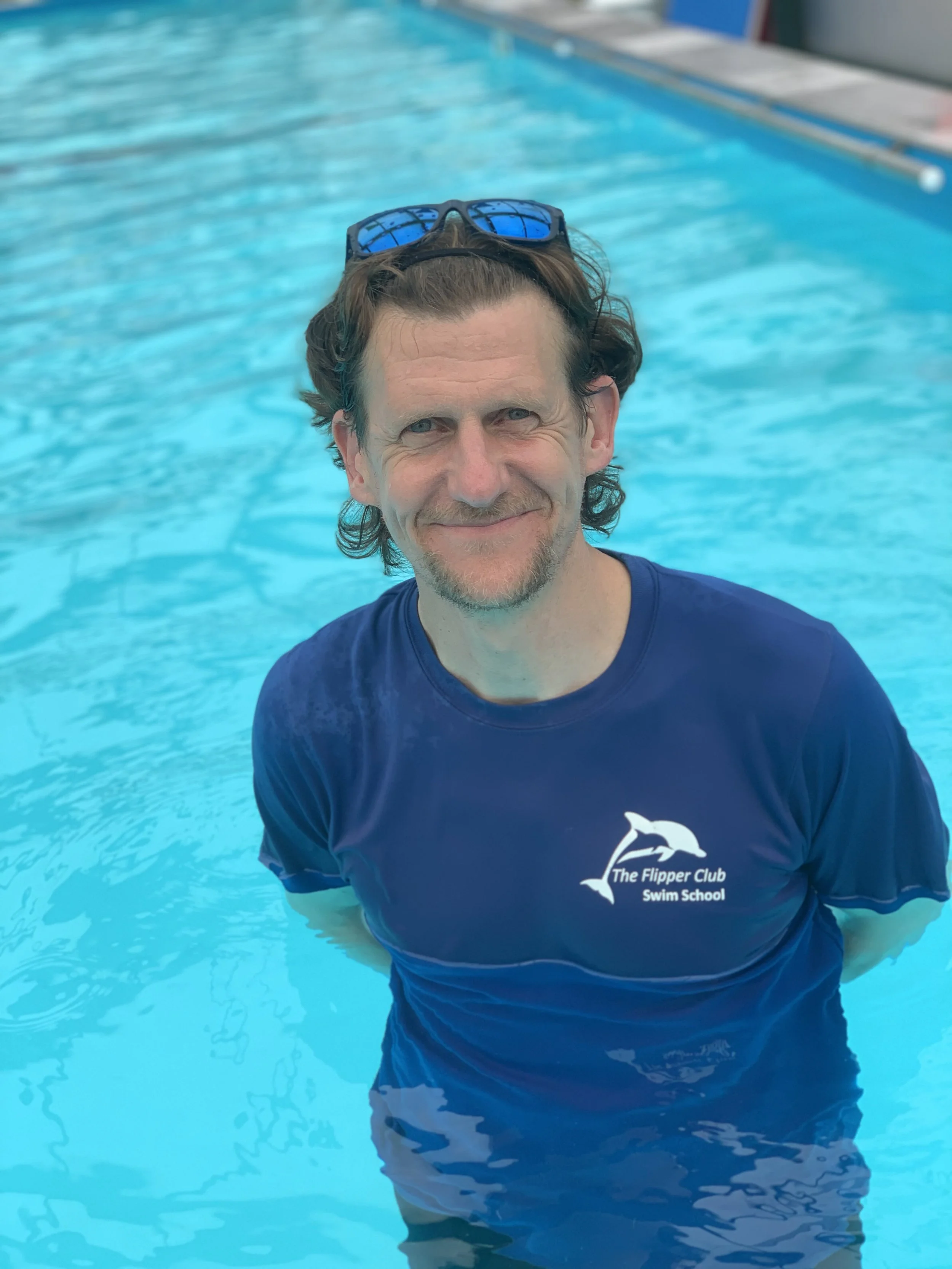 Man with glasses on head smiling in swimming pool, wearing a shirt with 'The Flipper Club Swim School' logo.
