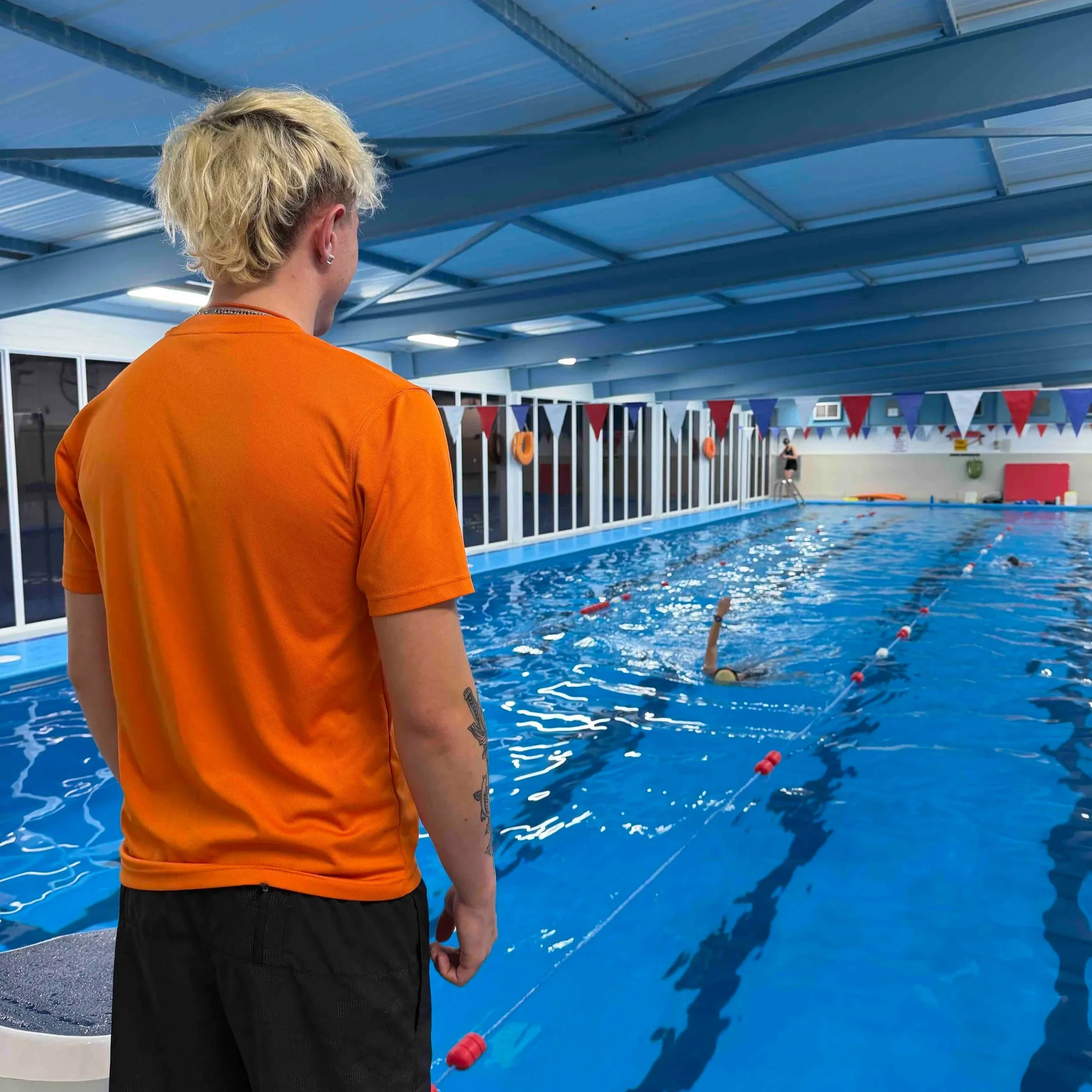 Flipper Club lifeguard wearing an orange shirt and black shorts standing by an indoor swimming pool, looking at the water, with a swimmer in the pool in the background. At Dunottar School swimming pool.
