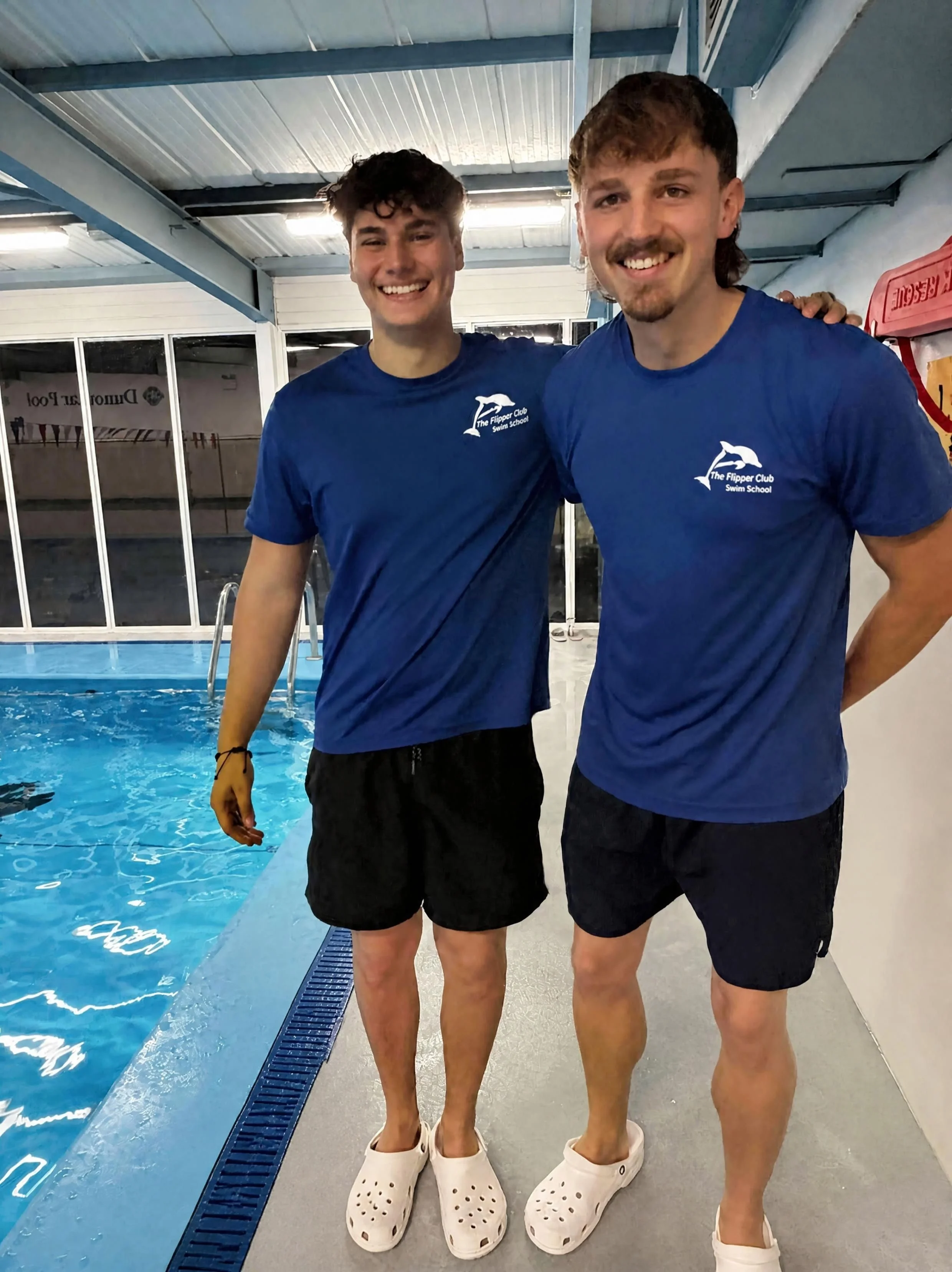 Two smiling young men in blue t-shirts and black shorts standing side by side by a swimming pool, wearing white Crocs shoes. The background shows swimming pool water, pool equipment, and a large window.