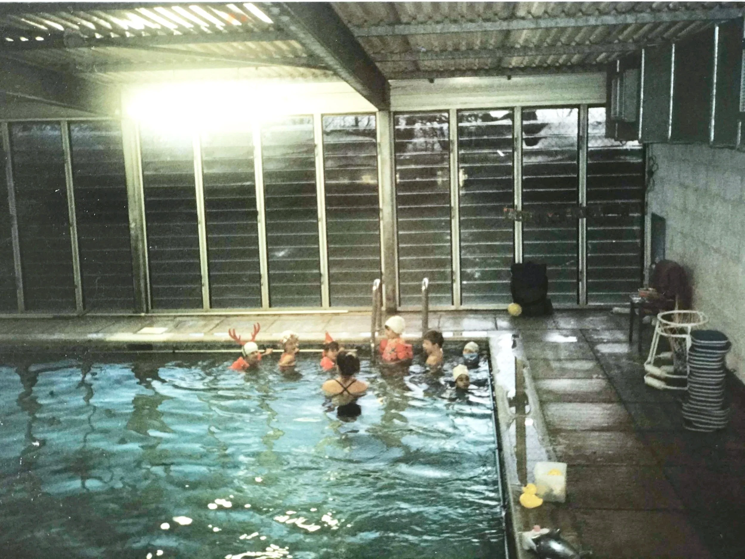 Indoor swimming pool with children in the water, some wearing swimming caps, and a number of pool toys and equipment along the edge.