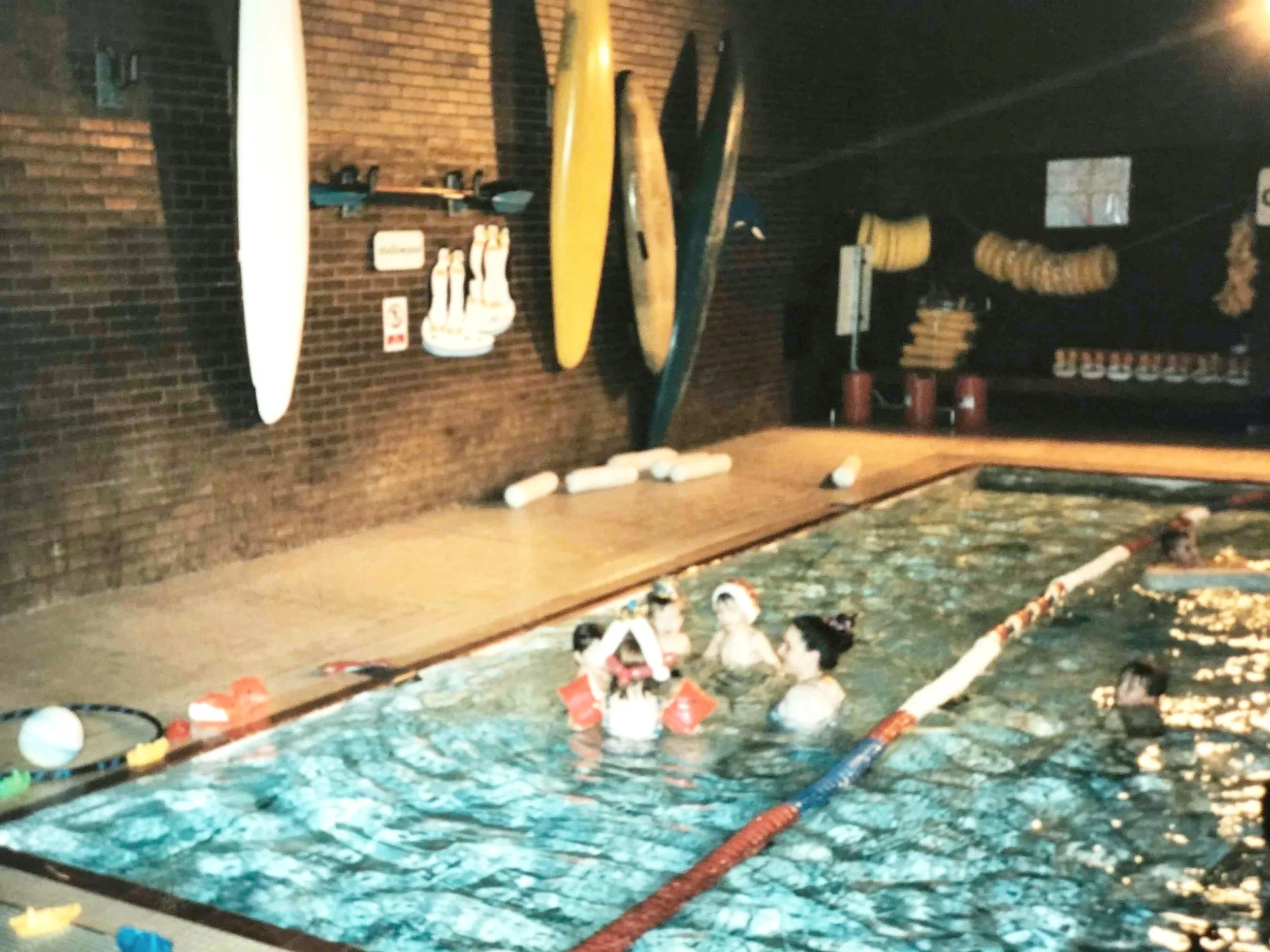 The image shows a swimming pool with water and floating toys, drinking straws, and other objects. In the background, there is a brick wall with surfboards and shelves holding bottles and other items, along with some hanging containers.