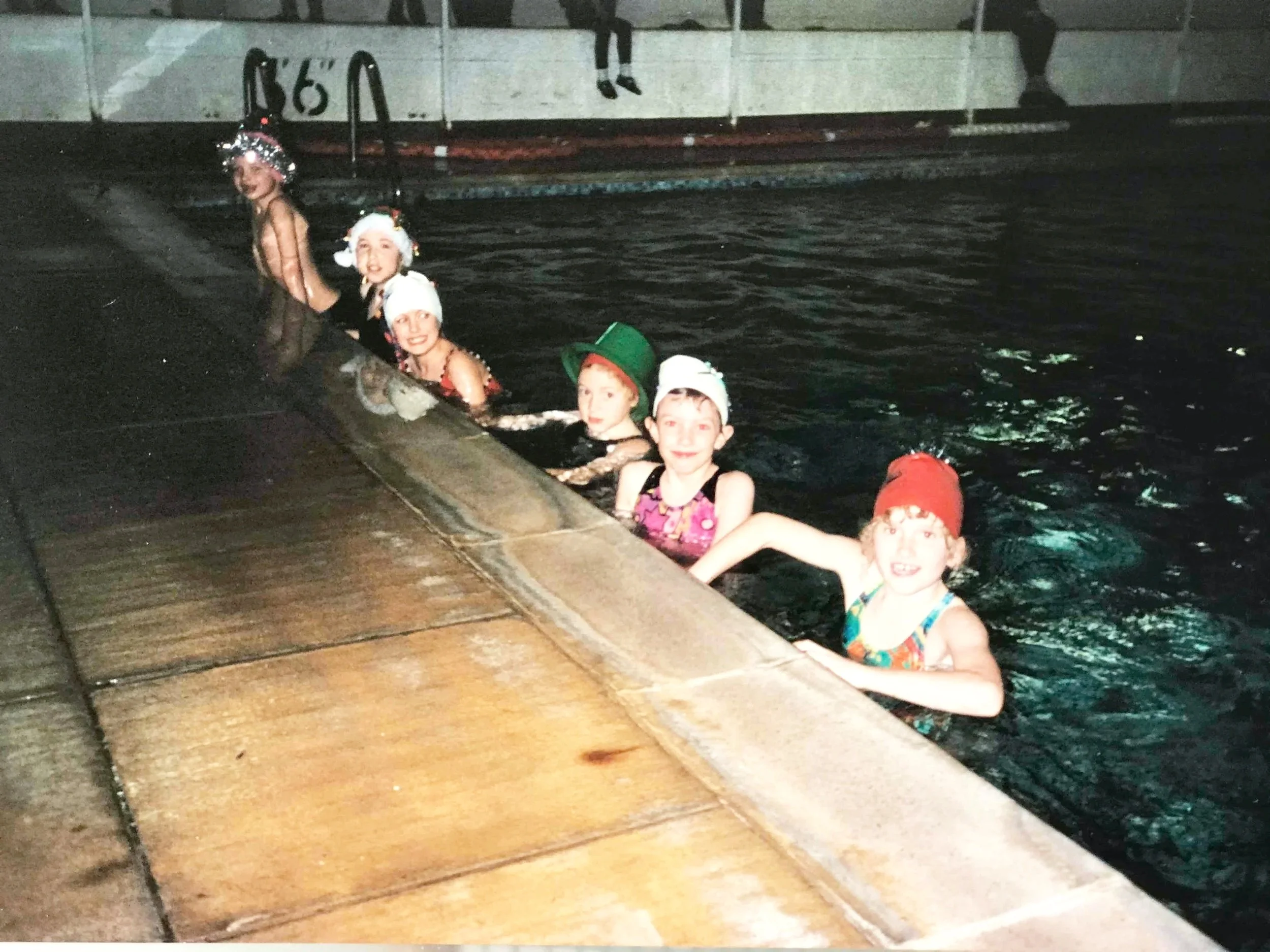 Children wearing festive holiday hats swimming in a pool at night, holding onto the edge of the pool.