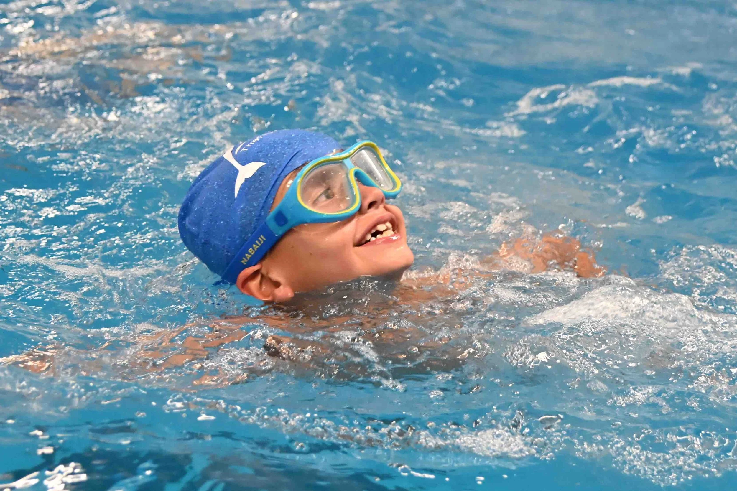 A boy swimming in a pool, wearing a blue swim cap and goggles, smiling and enjoying himself.