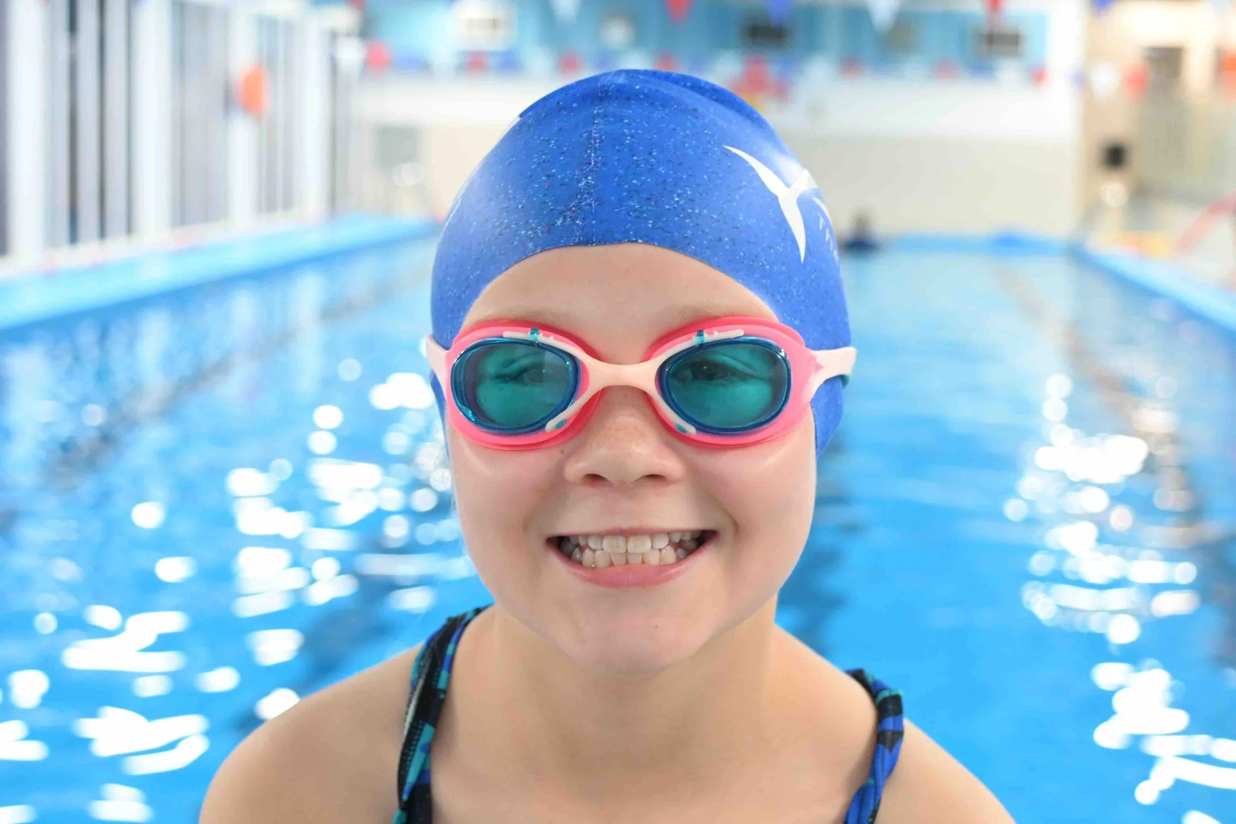 Close-up of a young girl in a blue swim cap and pink goggles smiling at the camera in an indoor swimming pool.