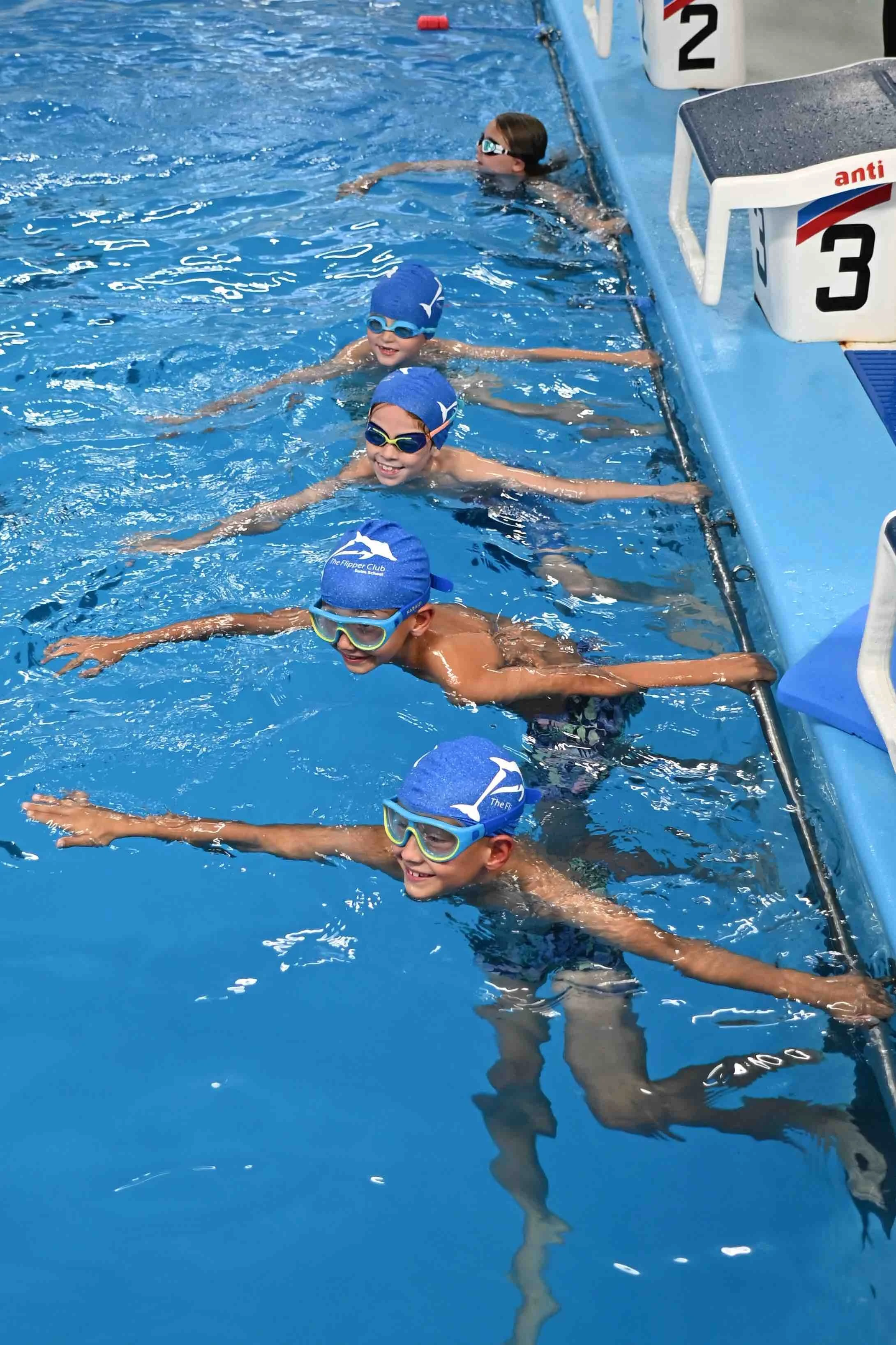 Kids in swim goggles and swim caps holding onto the side of a swimming pool after swimming practice or a race.