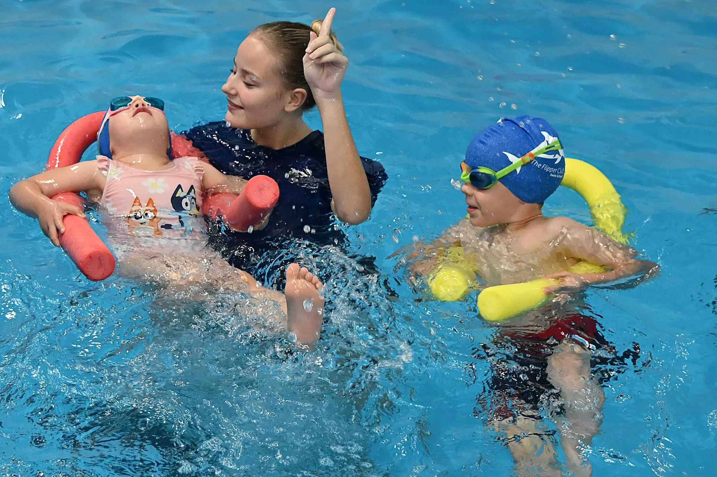 A young woman teaching two children to swim in a swimming pool. The children are wearing swimming goggles and floatation aids, while the woman is smiling and giving swimming instructions.