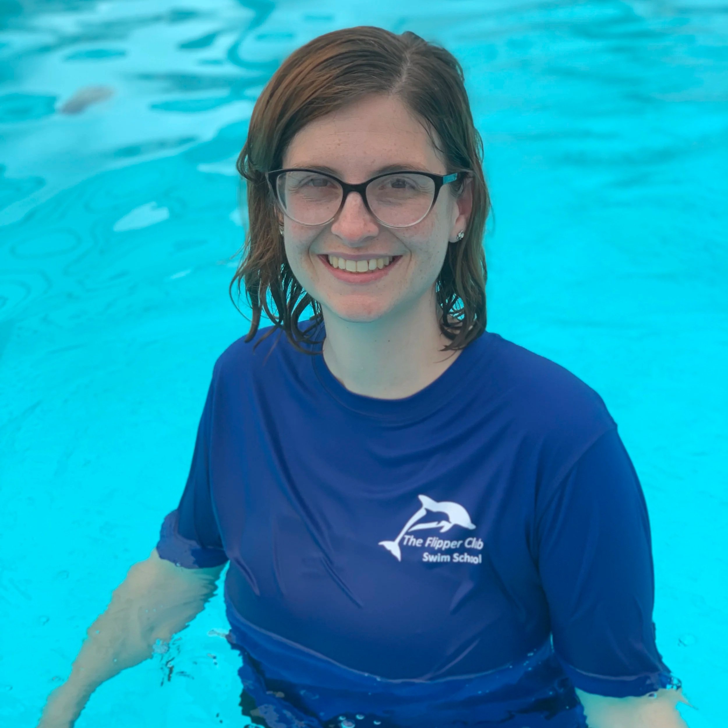 A woman in swimming goggles smiling in a swimming pool, wearing a blue swim shirt with the logo 'The Flipper Club Swim School'.