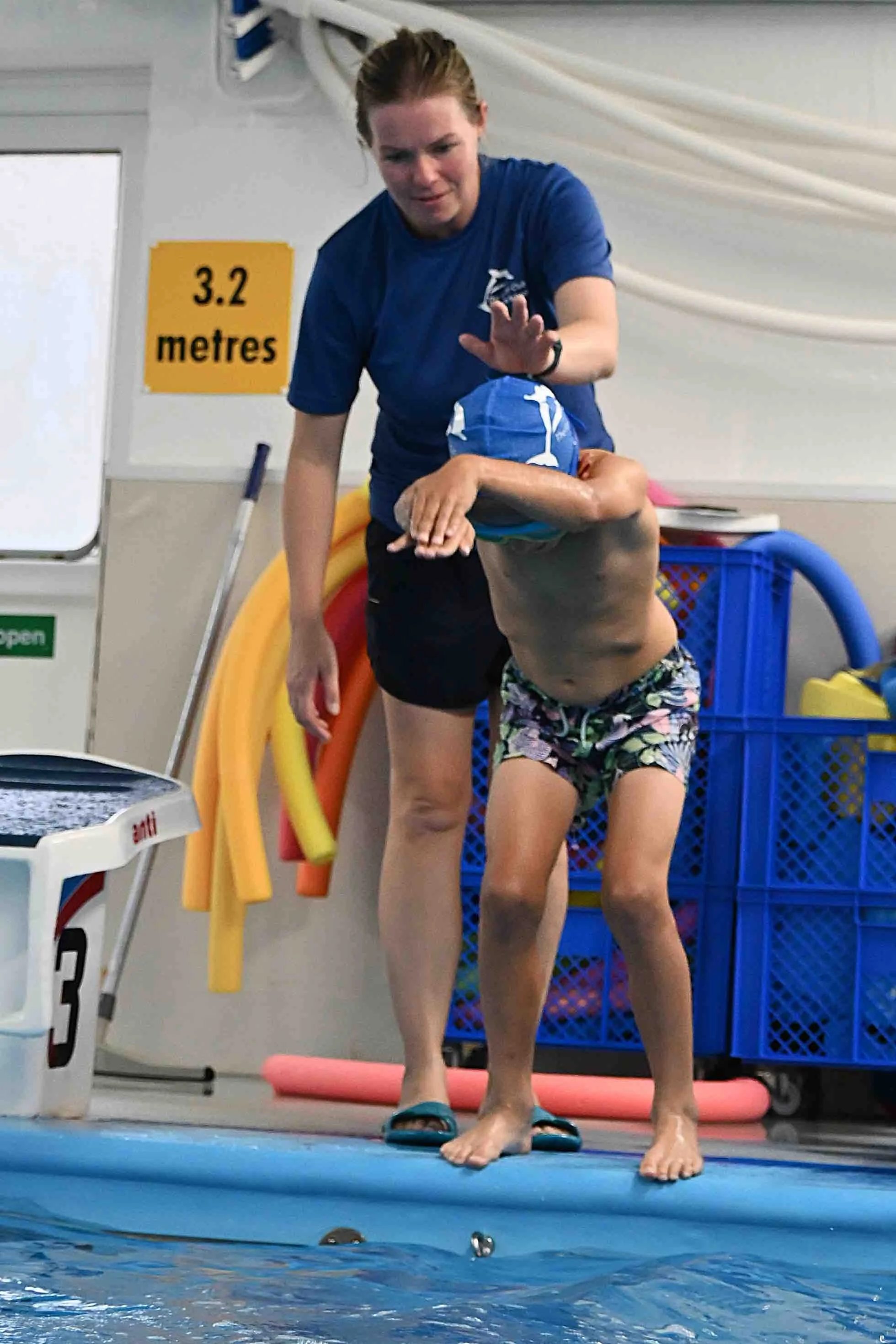 A woman is helping a child onto a swimming float at an indoor swimming pool. The woman is wearing a blue shirt and shorts, and the child is in a blue swim cap and patterned swim trunks. There are pool safety and equipment signs and items in the background.