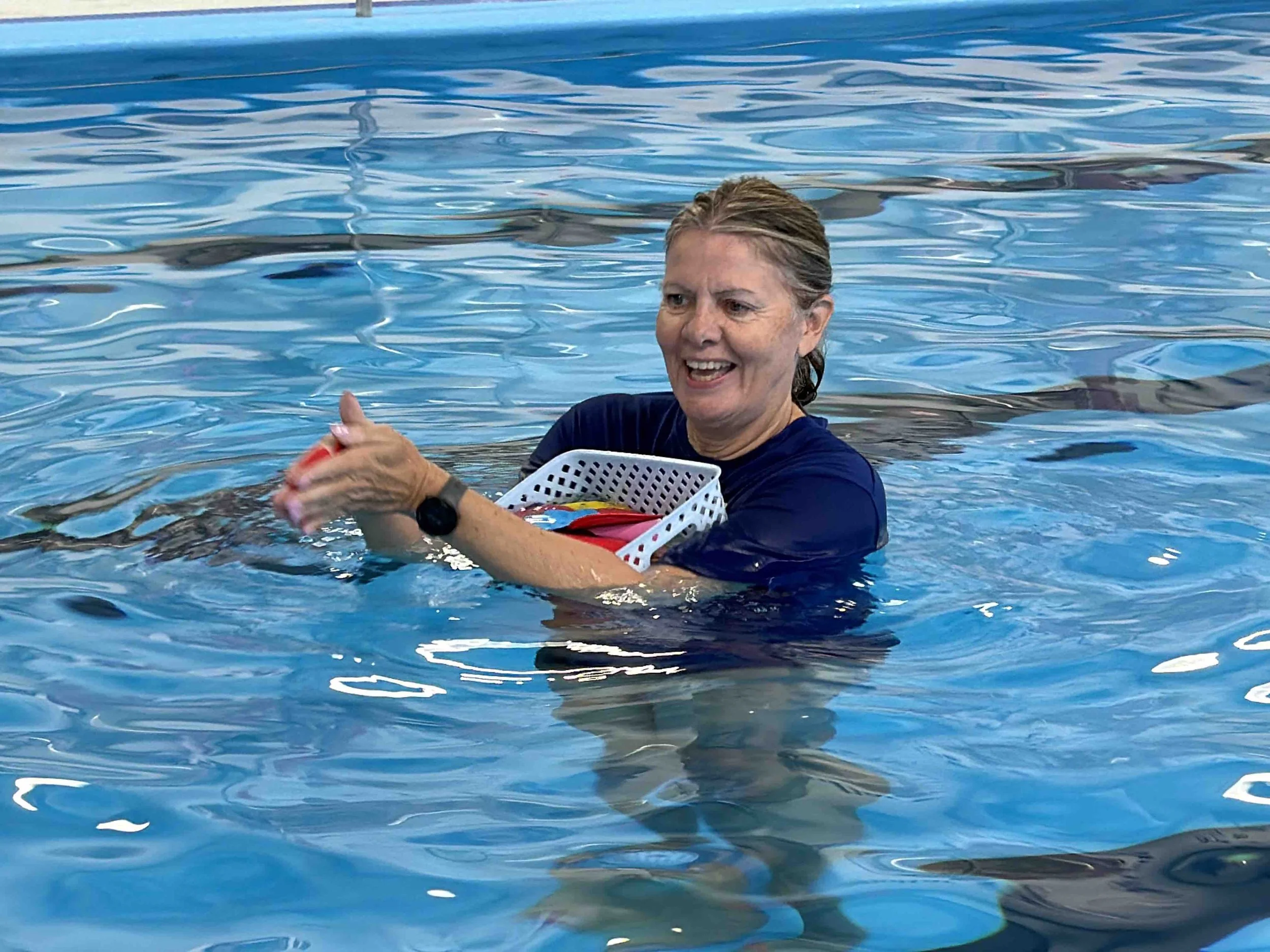 A woman in a navy shirt is smiling and holding a small red object while standing in a swimming pool. She is also holding a white basket with multicolored items inside. The background shows the water surface of the pool.