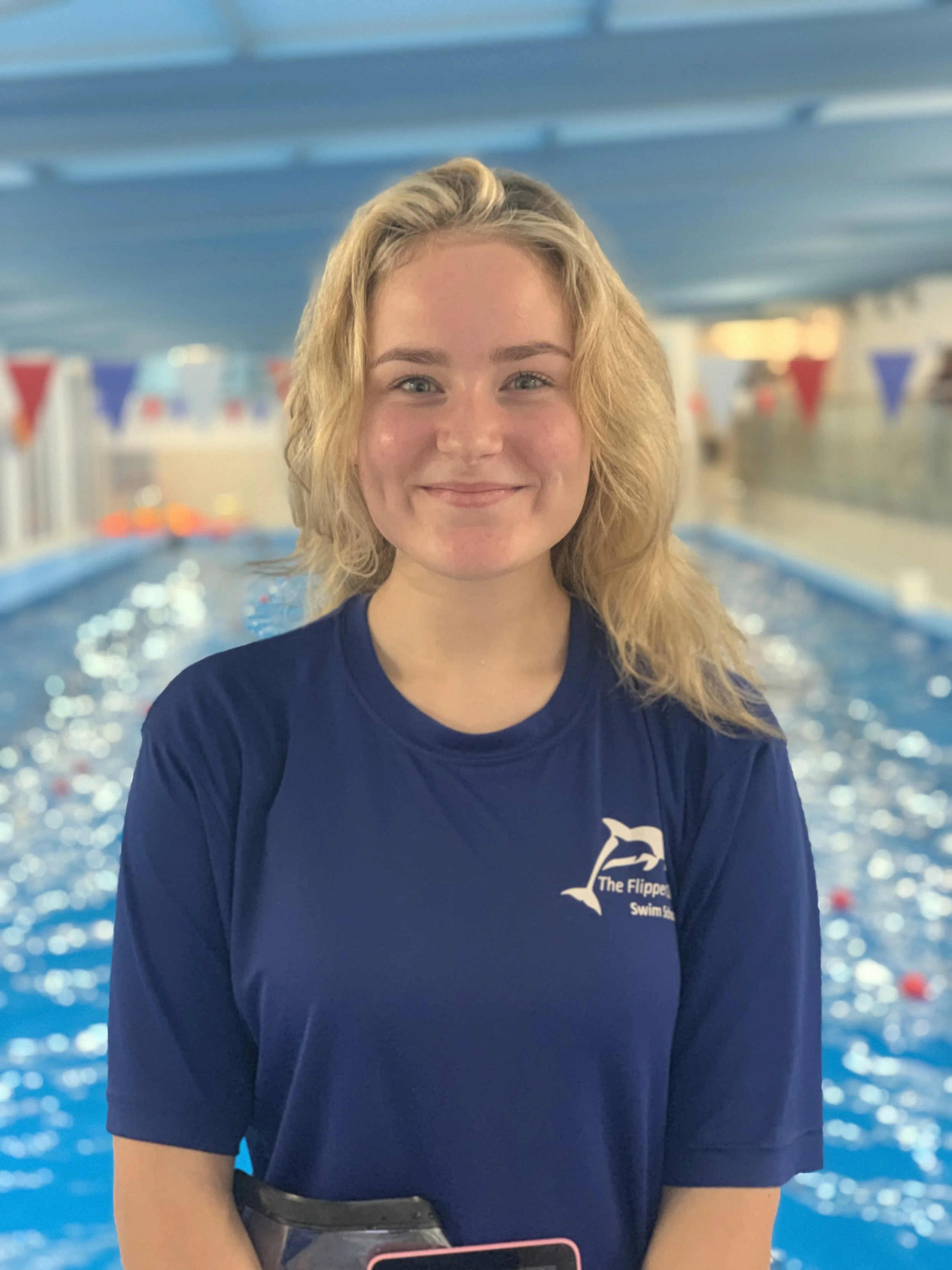 Close-up of a young woman with blonde hair smiling, standing in front of an indoor swimming pool with blue water and swimming lane dividers.