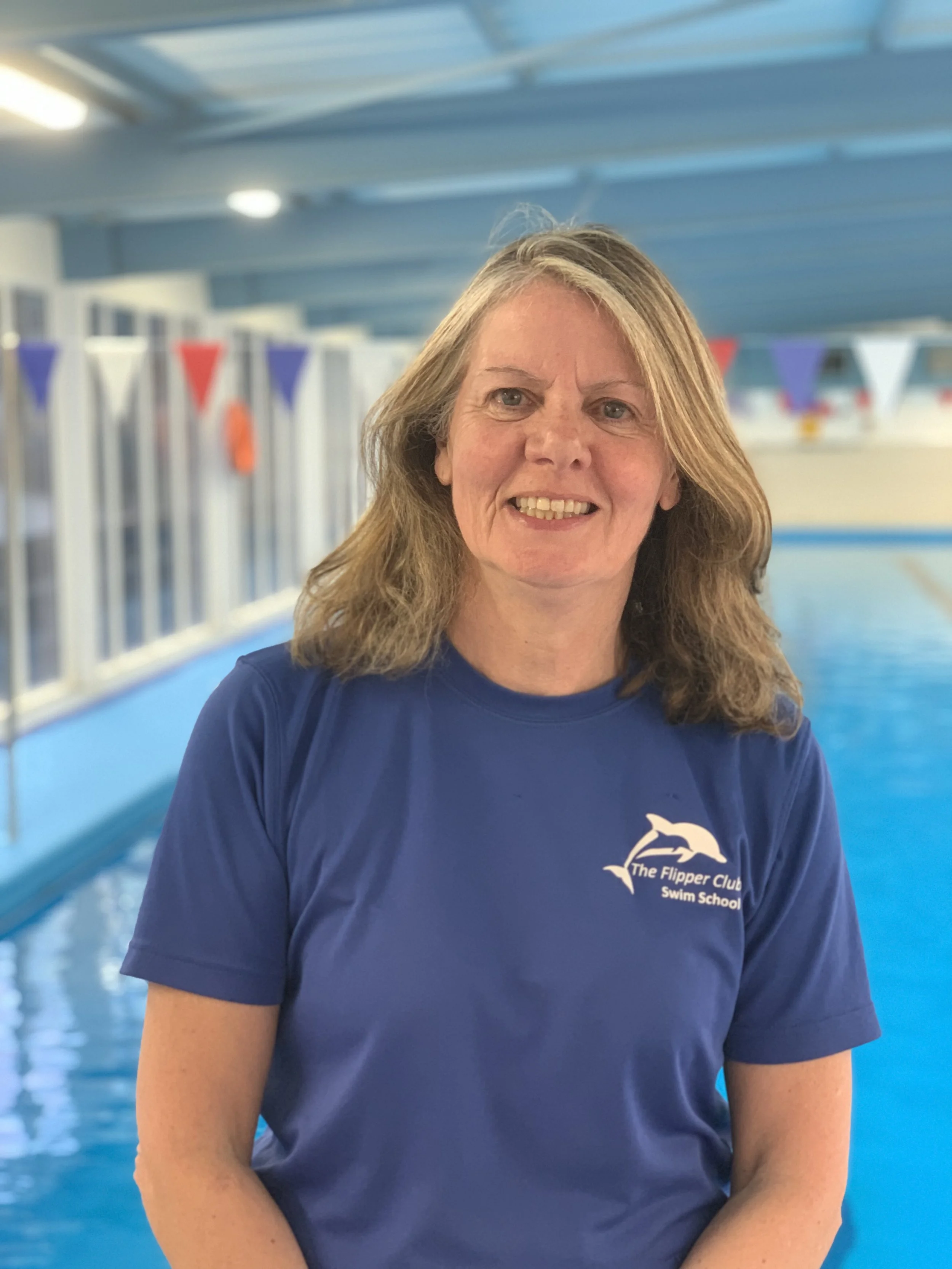 A woman with shoulder-length light brown hair smiling at the camera, wearing a blue t-shirt with a logo of dolphins and text that says 'The Flipper Club Swim School,' standing in front of a swimming pool with blue water and flags hanging in the background.