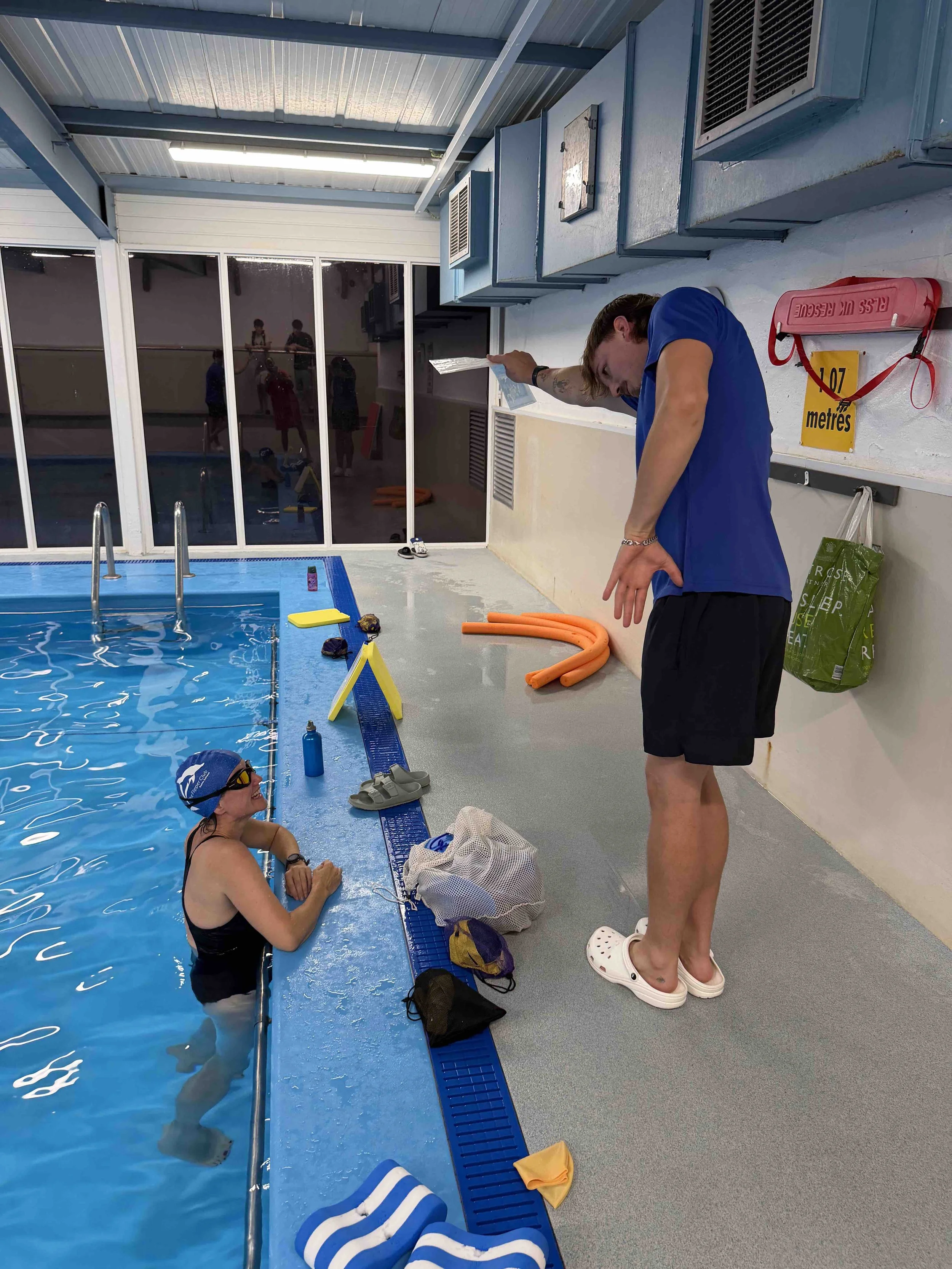 A swimming pool area with two people in the pool and one person standing outside the pool. The person in the pool is wearing a blue swim cap and goggles, and the person on the poolside is holding a clipboard. Various equipment and accessories are on the floor, including a towel, a bag, and flotation devices. The background shows large windows with reflections of other people and a brightly lit indoor space.
