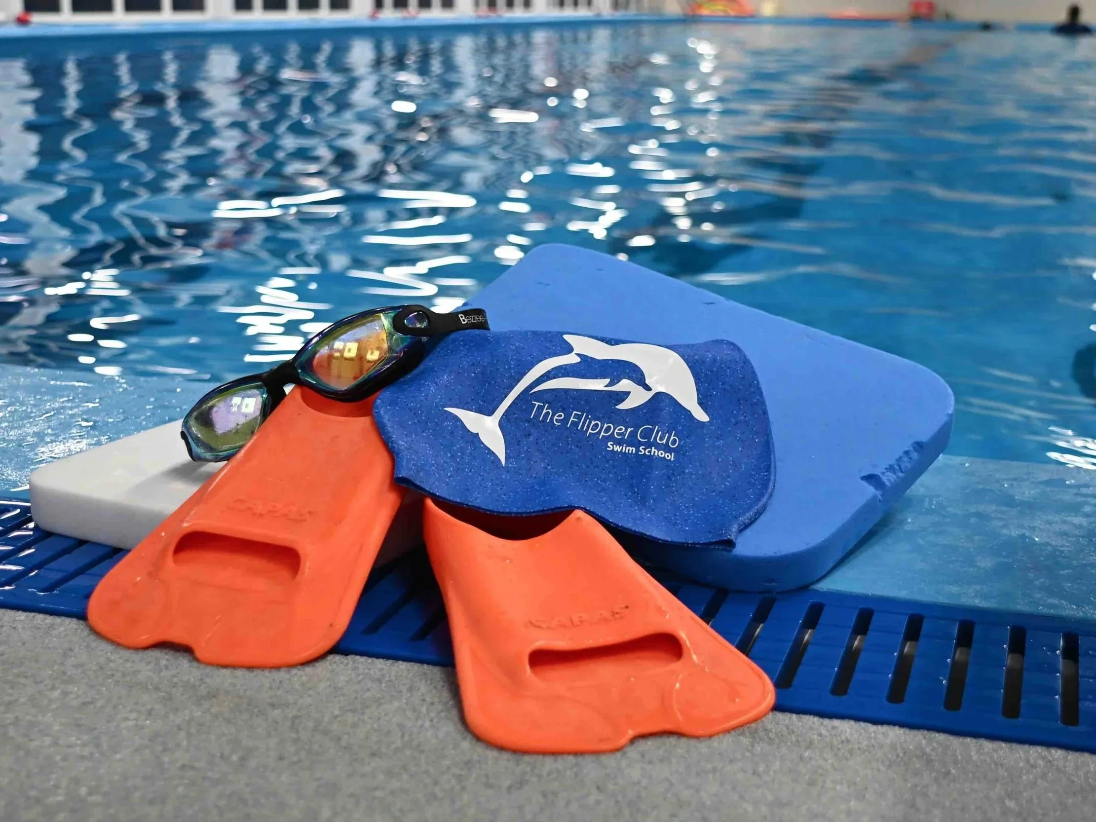 Swimming goggles, orange swim fins, blue swim cap with dolphin logo and the text 'The Flipper Club Swim School,' and a pair of goggles, all arranged on a blue pool deck with Dunottar swimming pool in the background.