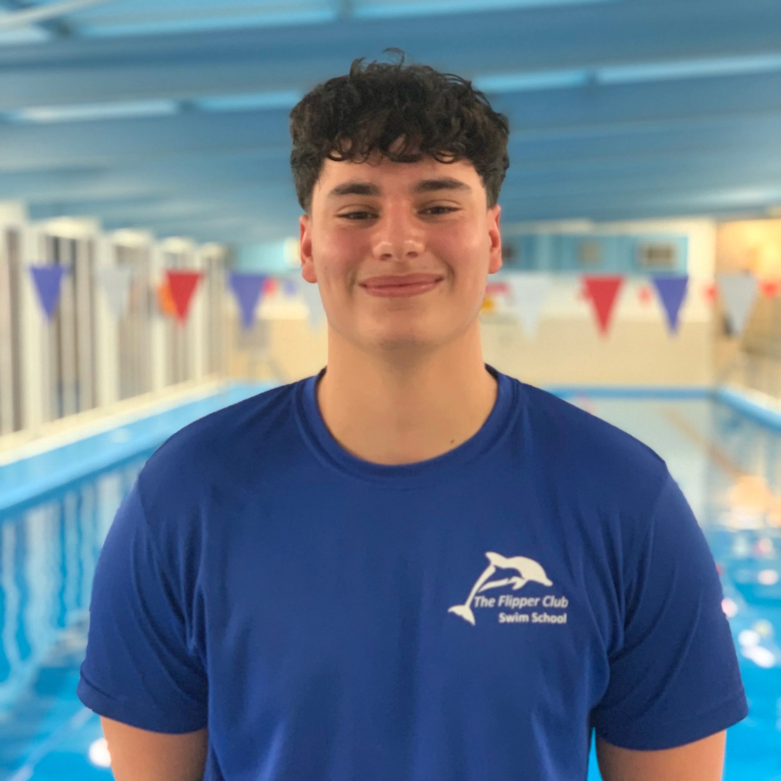 Close-up of a young man's smiling face wearing a blue swim shirt with the text 'The Flipper Club Swim School' on it, in a swimming pool area with blue lane dividers and flags in the background.
