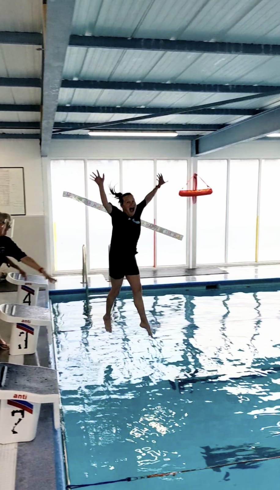 A woman jumping into a swimming pool with her arms raised and a wide smile, wearing a black shirt and shorts, with a measuring tape around her neck, inside an indoor swimming facility.