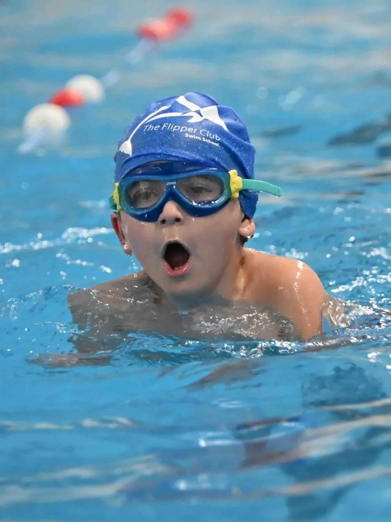 A young boy swimming in a pool with a blue swim cap and goggles, with his mouth open and an expression of surprise or excitement.