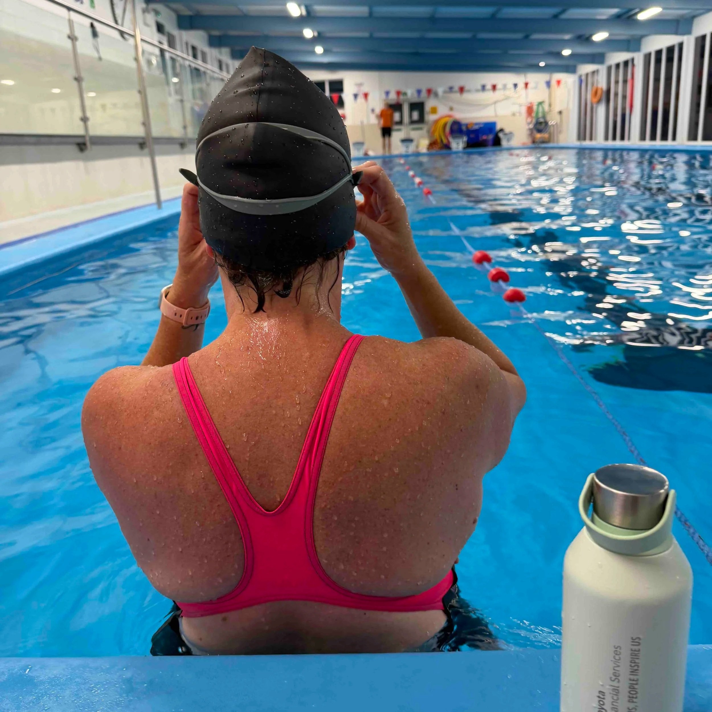A person wearing a black swim cap and pink swimsuit is preparing to swim in an indoor swimming pool. The person is holding the swim cap, water droplets are visible on their skin and cap. The pool has blue water and red and white lane dividers. The background shows the pool's indoor facilities with a person standing near the far end.