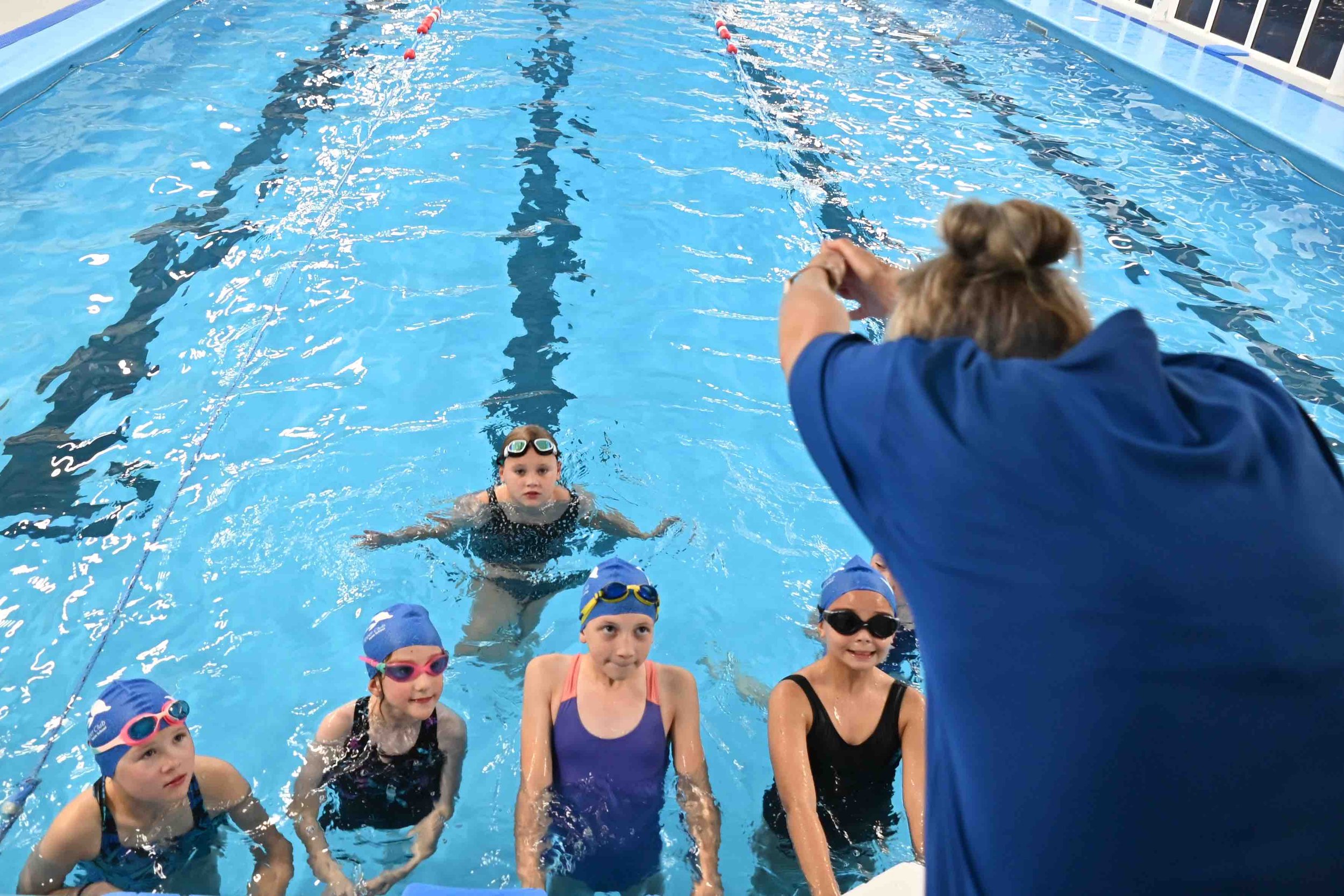 A group of children in swim gear standing in a swimming pool. An adult is leaning over to talk to them. The kids are wearing swimming caps and goggles.