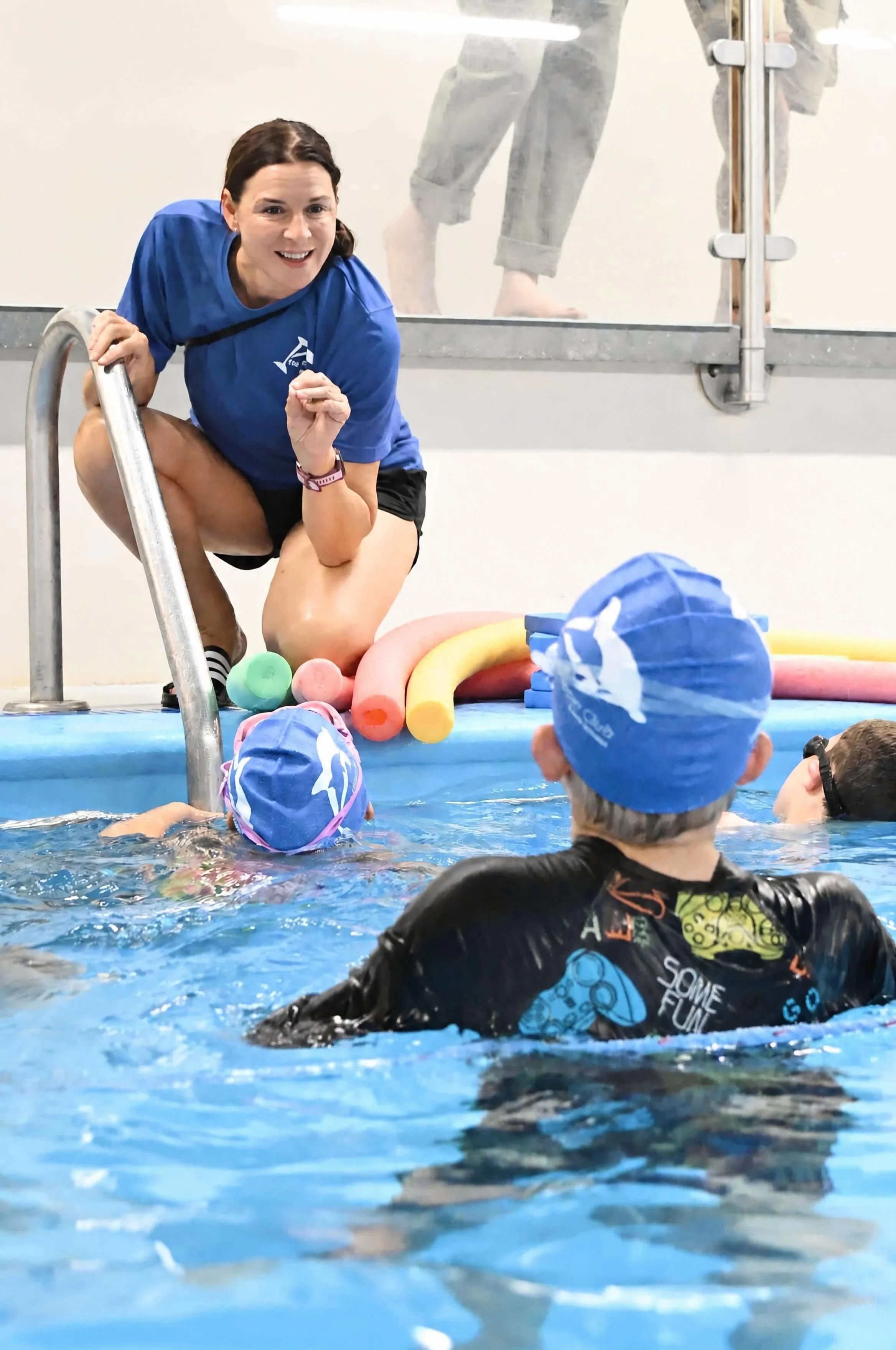A woman kneeling at the edge of a swimming pool talking to a group of children wearing swim caps. The woman is in a blue shirt, and the children are in colorful swimwear and caps. Foam pool noodles are on the pool deck.