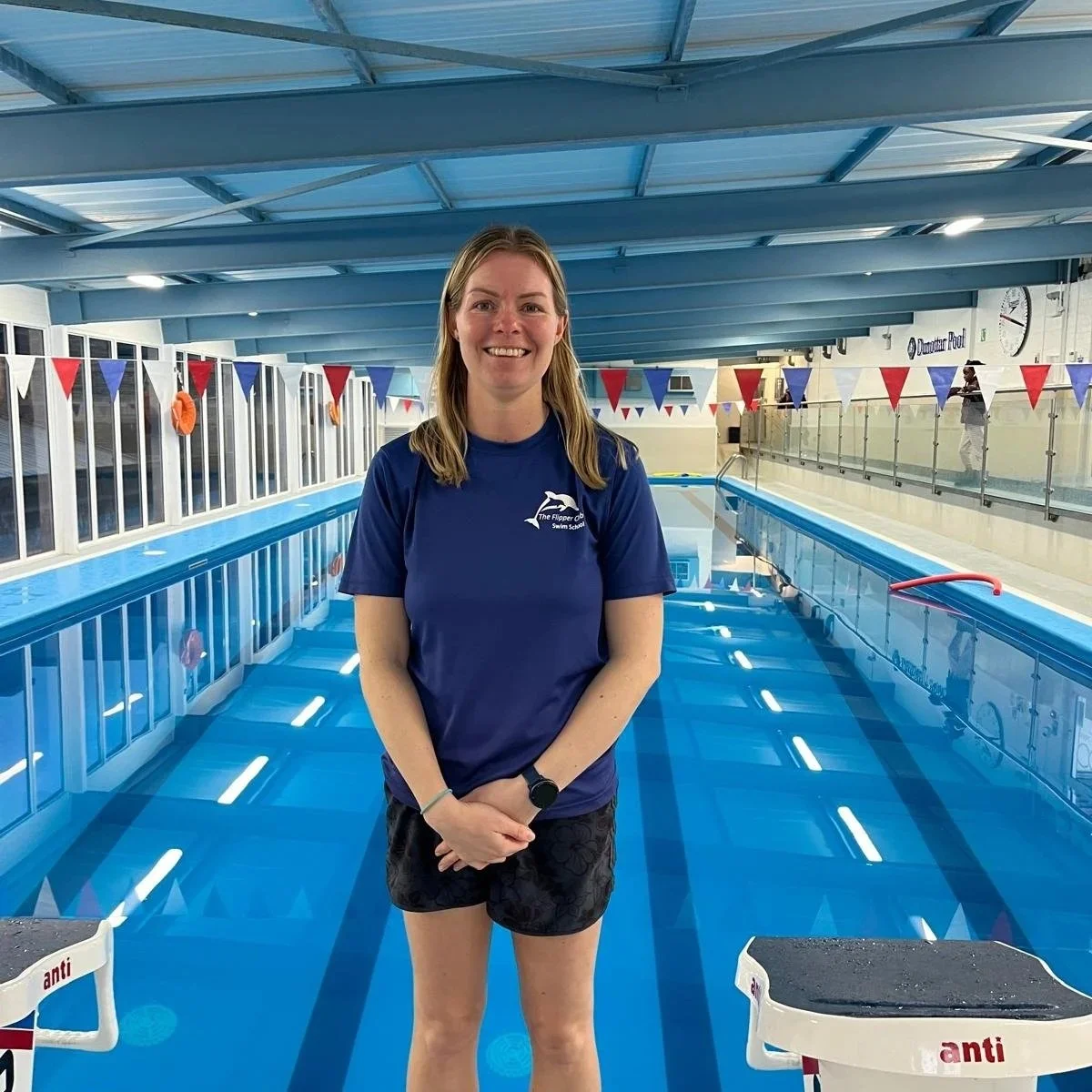 A woman standing in front of an indoor swimming pool, wearing a blue shirt with a dolphin logo and black shorts, smiling at the camera.