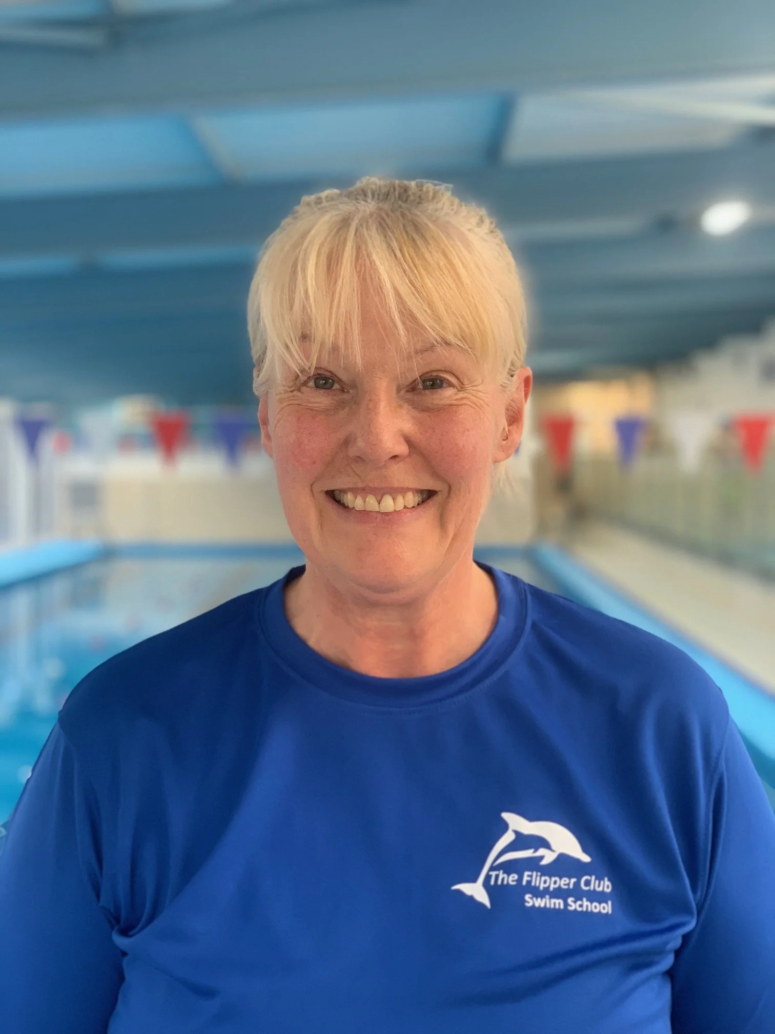 A smiling woman with short blonde hair in a blue swim team uniform standing in an indoor swimming pool. The logo on her shirt reads 'The Flipper Club Swim School' and features a dolphin graphic. In the background, there are blue and red triangular banners hanging along the pool area.