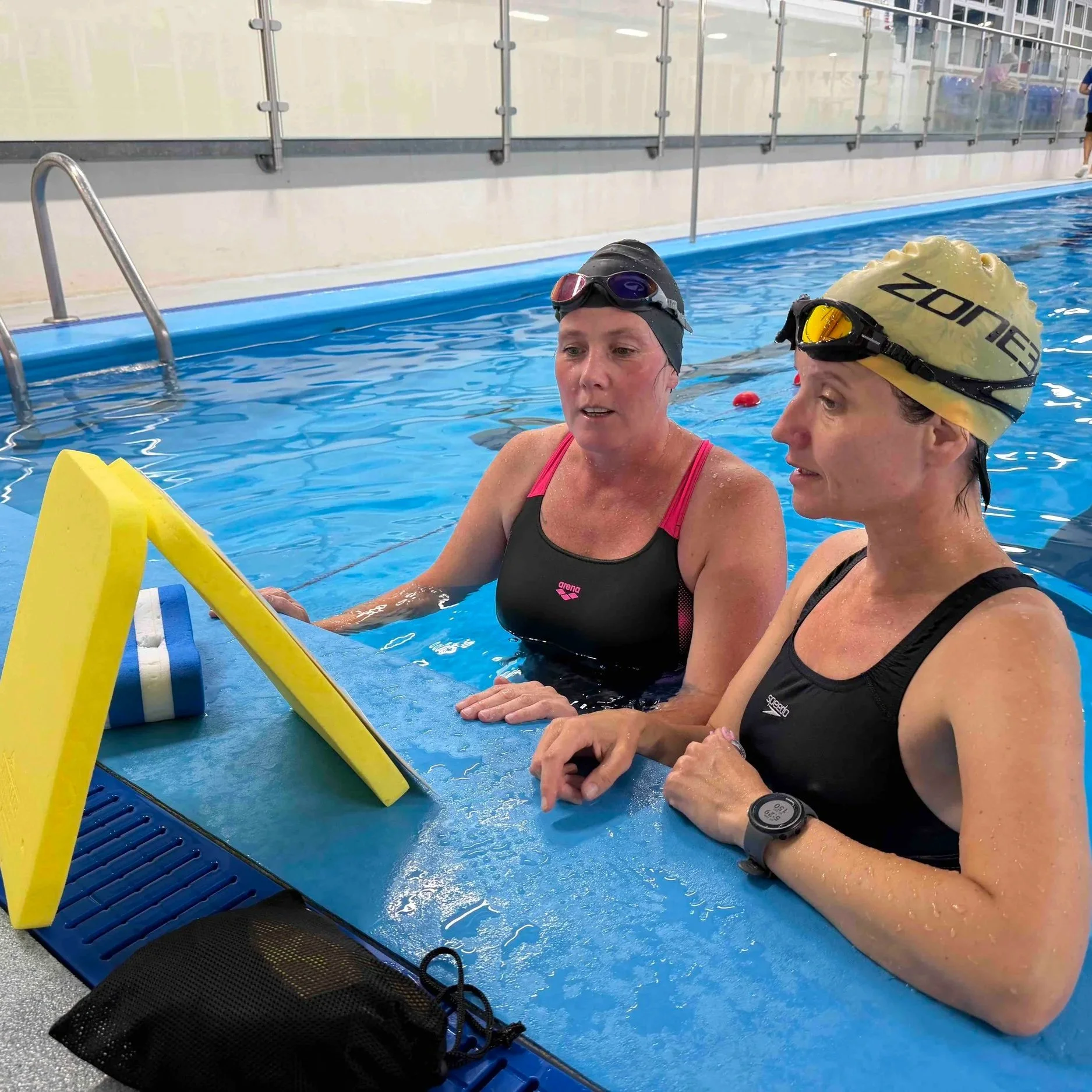 Two women in swimwear and swim caps in an indoor swimming pool, engaged in conversation, with a yellow pool noodle and swimming equipment nearby.