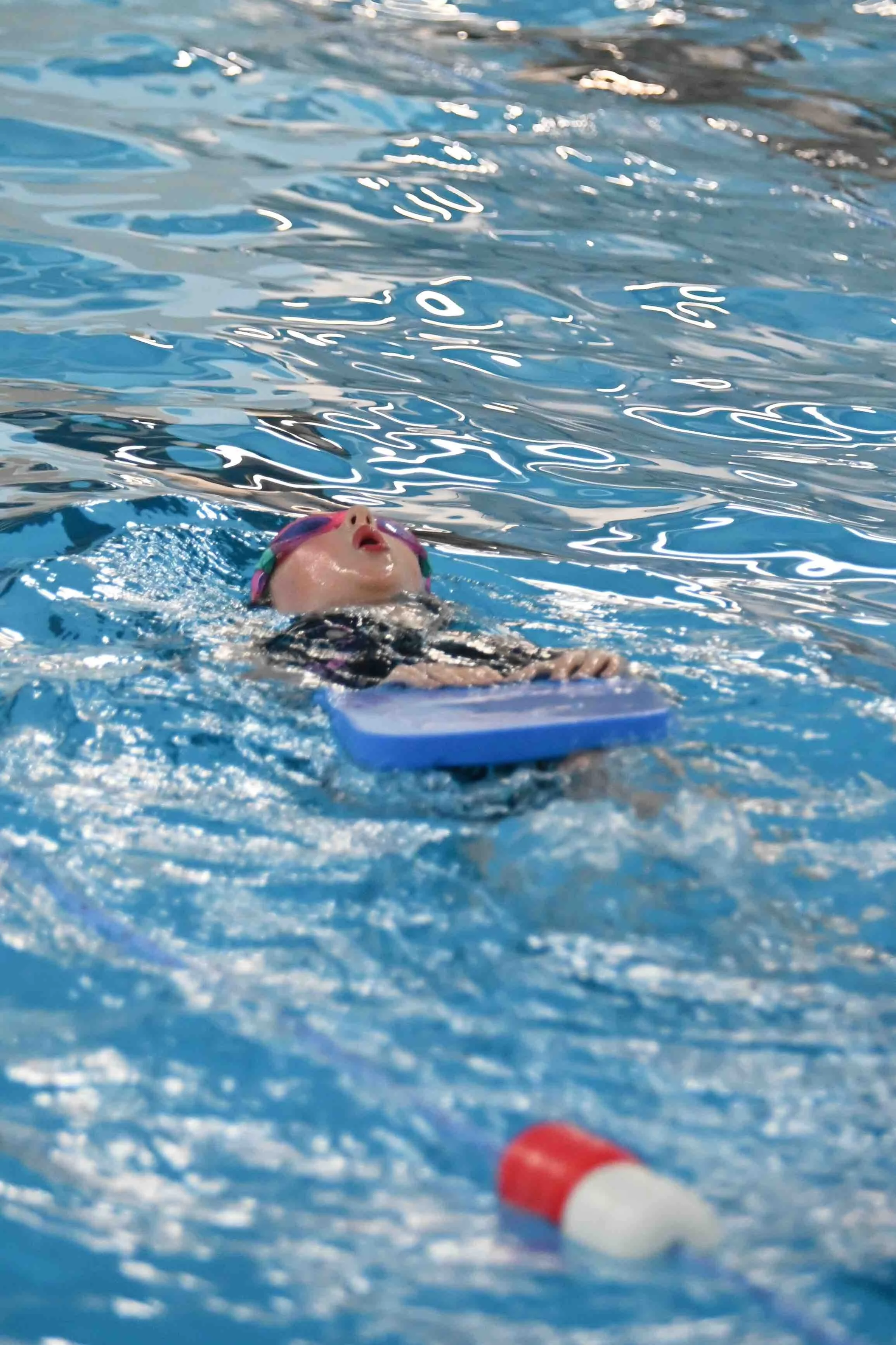 Swimmer with pink cap and goggles in a swimming pool, holding onto a blue kickboard, with a red and white buoy in the water nearby.