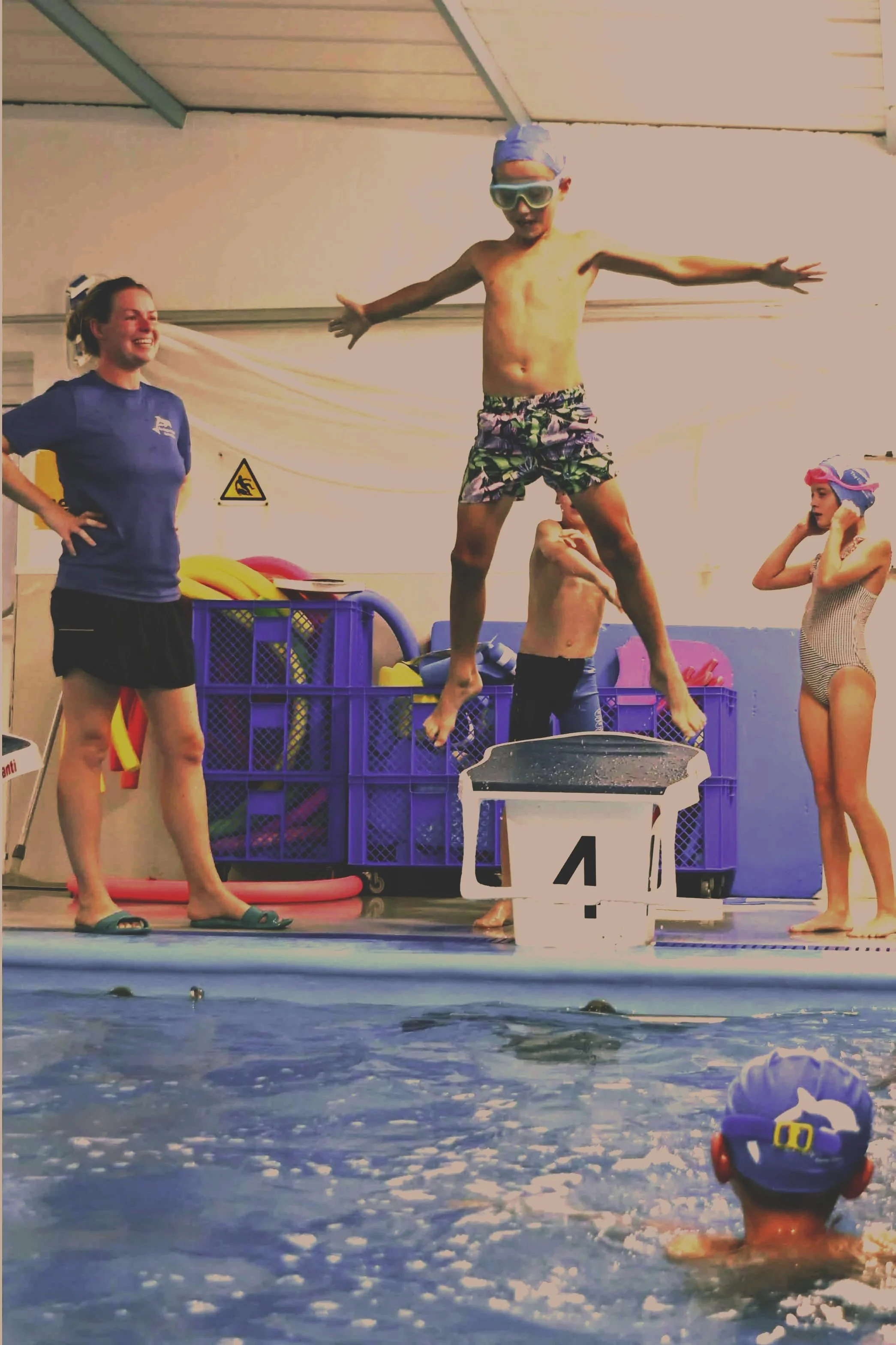 Children and an adult at an indoor swimming pool. A boy in swim shorts and goggles stands on a platform with arms outstretched. A girl in a striped swimsuit and swimming cap stands nearby, adjusting her goggles. An adult woman in a blue shirt and black shorts stands with hands on her hips, smiling. Swimming equipment is visible in the background.