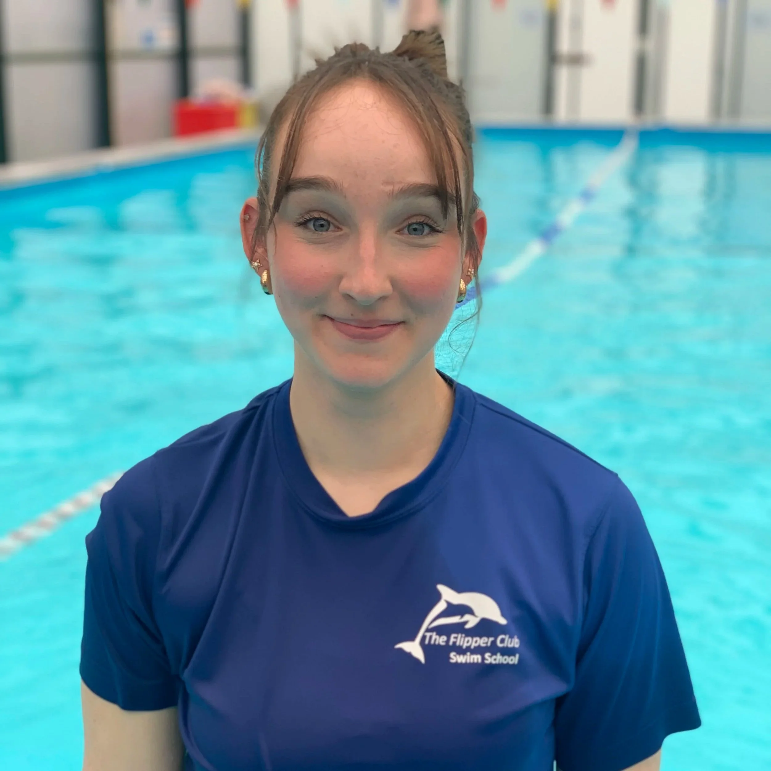 Close-up of a smiling girl in a blue T-shirt with the logo of The Flipper Club Swim School, standing in a swimming pool with lane lines visible in the background.