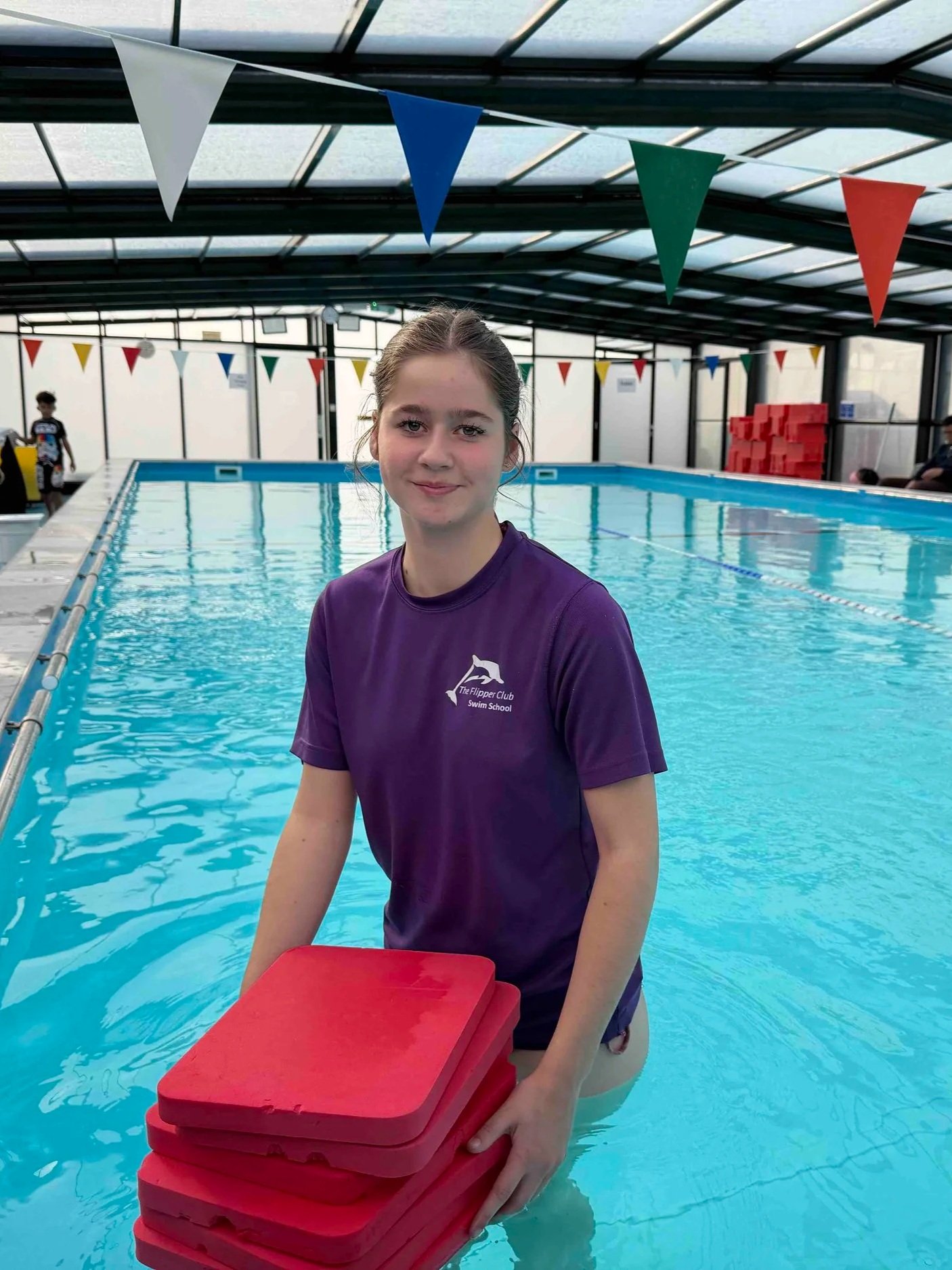 A young girl standing in a swimming pool holding a stack of red tiles or trays. She is wearing a purple t-shirt with a logo that reads 'The Flipper Club Swim School.' The indoor pool area has colorful pennant banners hanging from the ceiling, and there are other people in the background, some near the poolside.