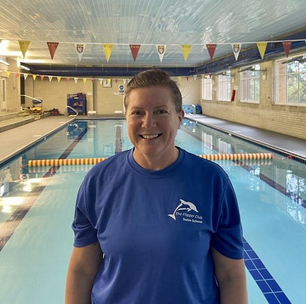 A woman standing in front of an indoor swimming pool, wearing a blue polo shirt with a swim club logo, smiling at the camera.