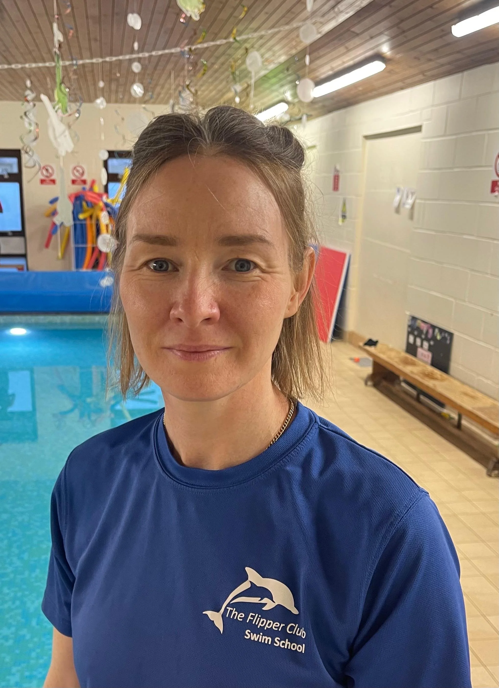 A woman in a blue shirt with the logo of The Flipper Club Swim School stands in an indoor swimming pool area. The background features hanging decorations, a wooden ceiling, fluorescent lighting, and a bench along the wall.