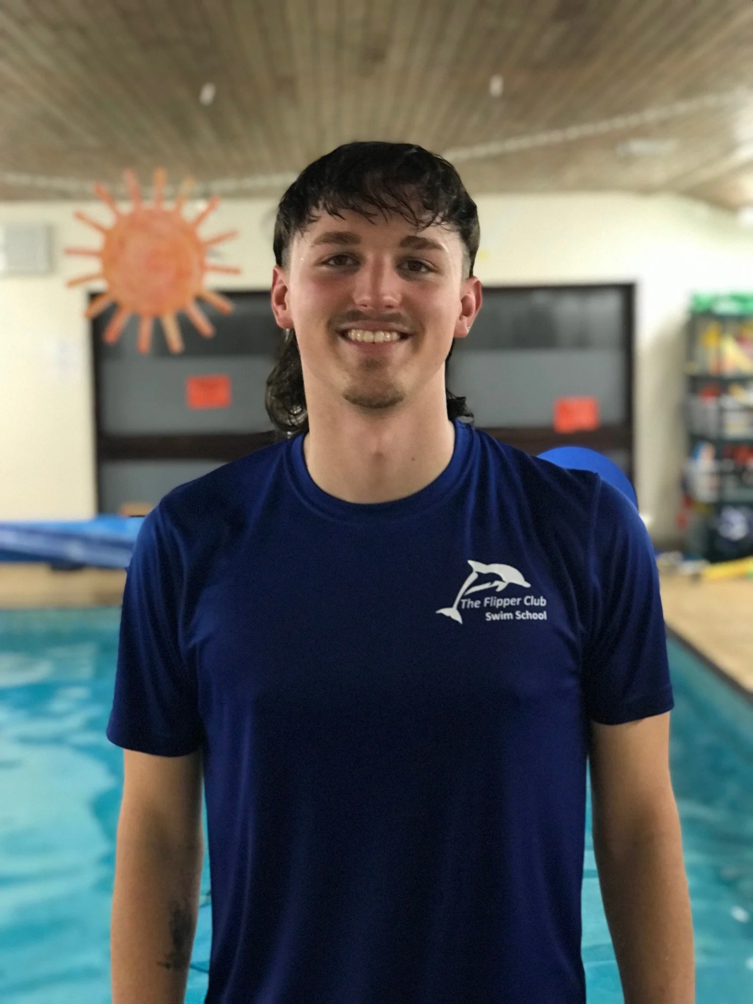 A young man with dark hair, smiling, standing near a swimming pool. He is wearing a blue t-shirt with a dolphin logo and the text 'The Flipper Club Swim School.' In the background, there is a mural of an orange sun on the wall and a bookshelf filled with books.