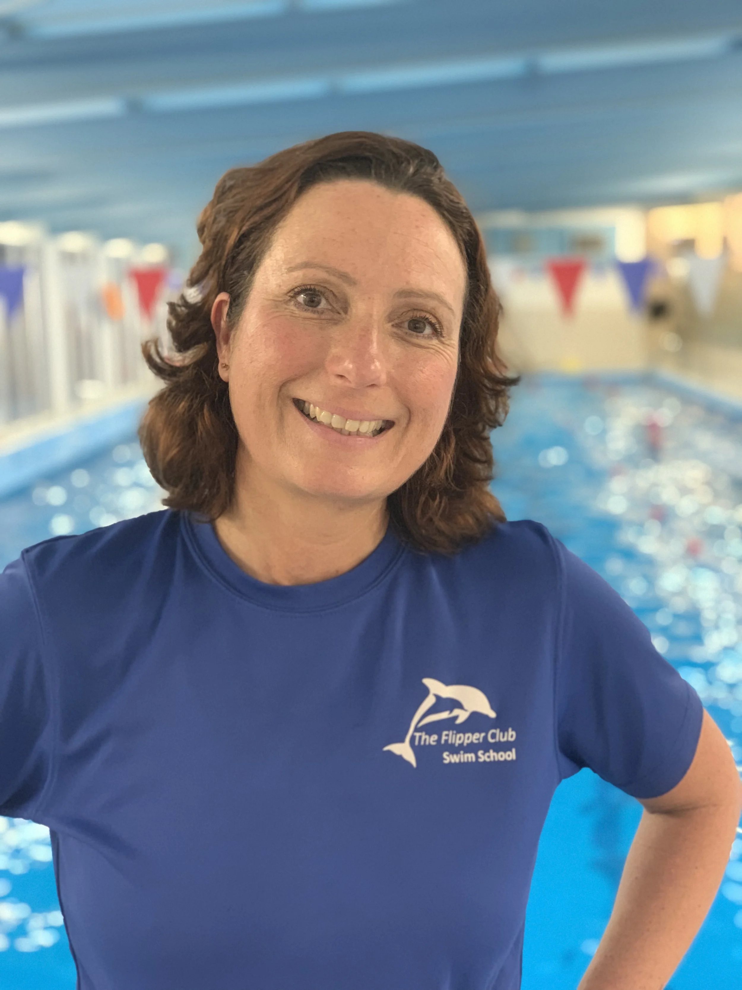 A smiling woman with curly brown hair at an indoor swimming pool, wearing a blue swim shirt with a dolphin logo and the text 'The Flipper Club Swim School'.