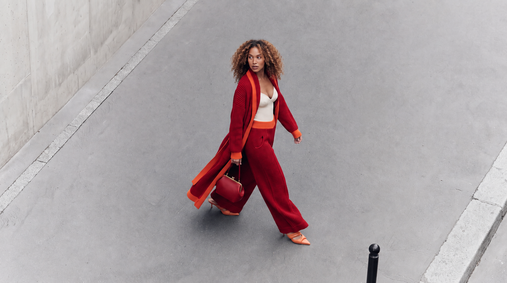 A woman walking on a concrete sidewalk next to a building wall, carrying a red handbag, wearing a white top, red and orange layered coat, red pants, and peach-colored high heels, with curly hair.