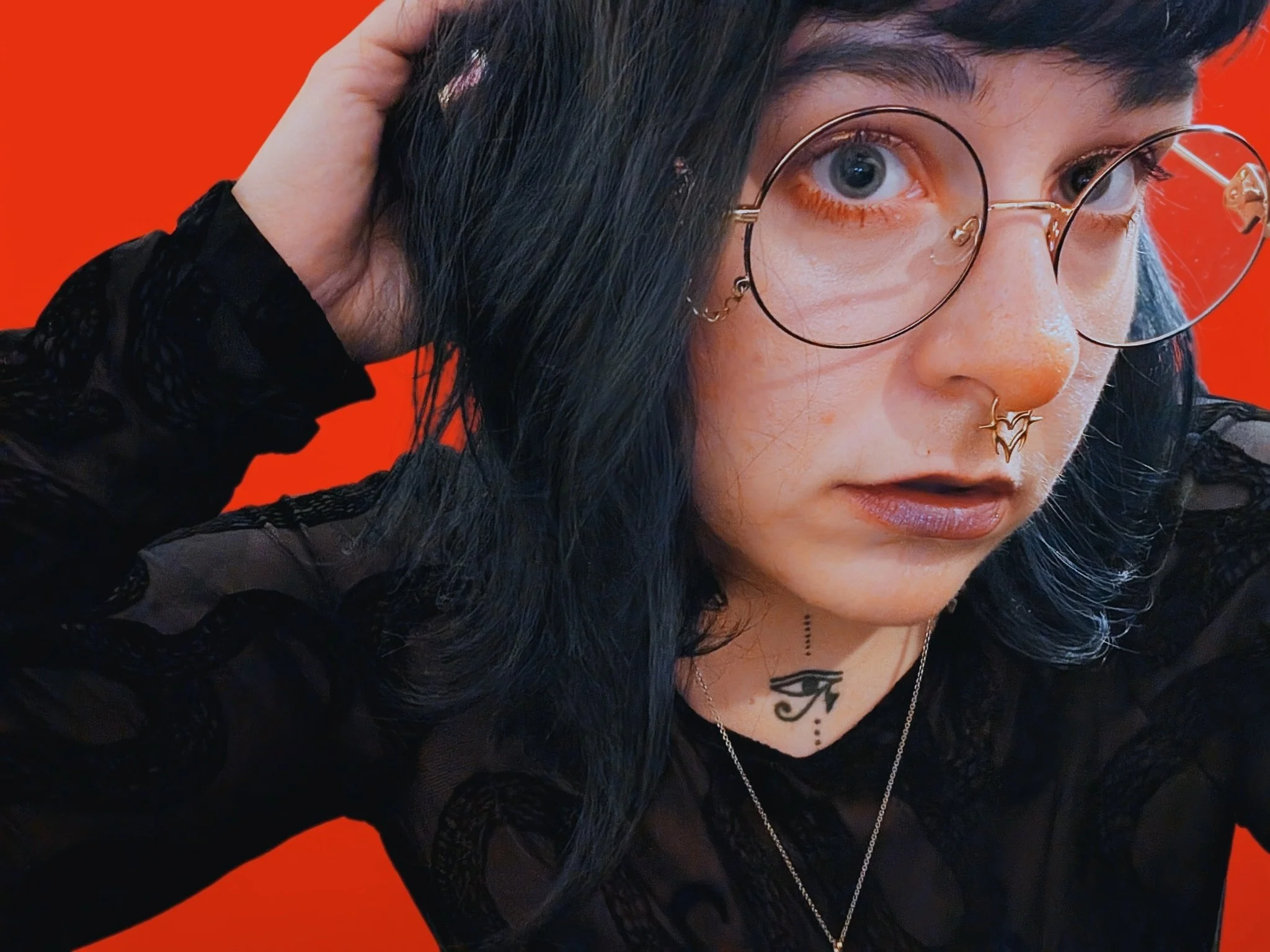 Close-up of a young person with dark hair, wearing large round glasses, facial piercings, and black lace clothing, against a bright red background.