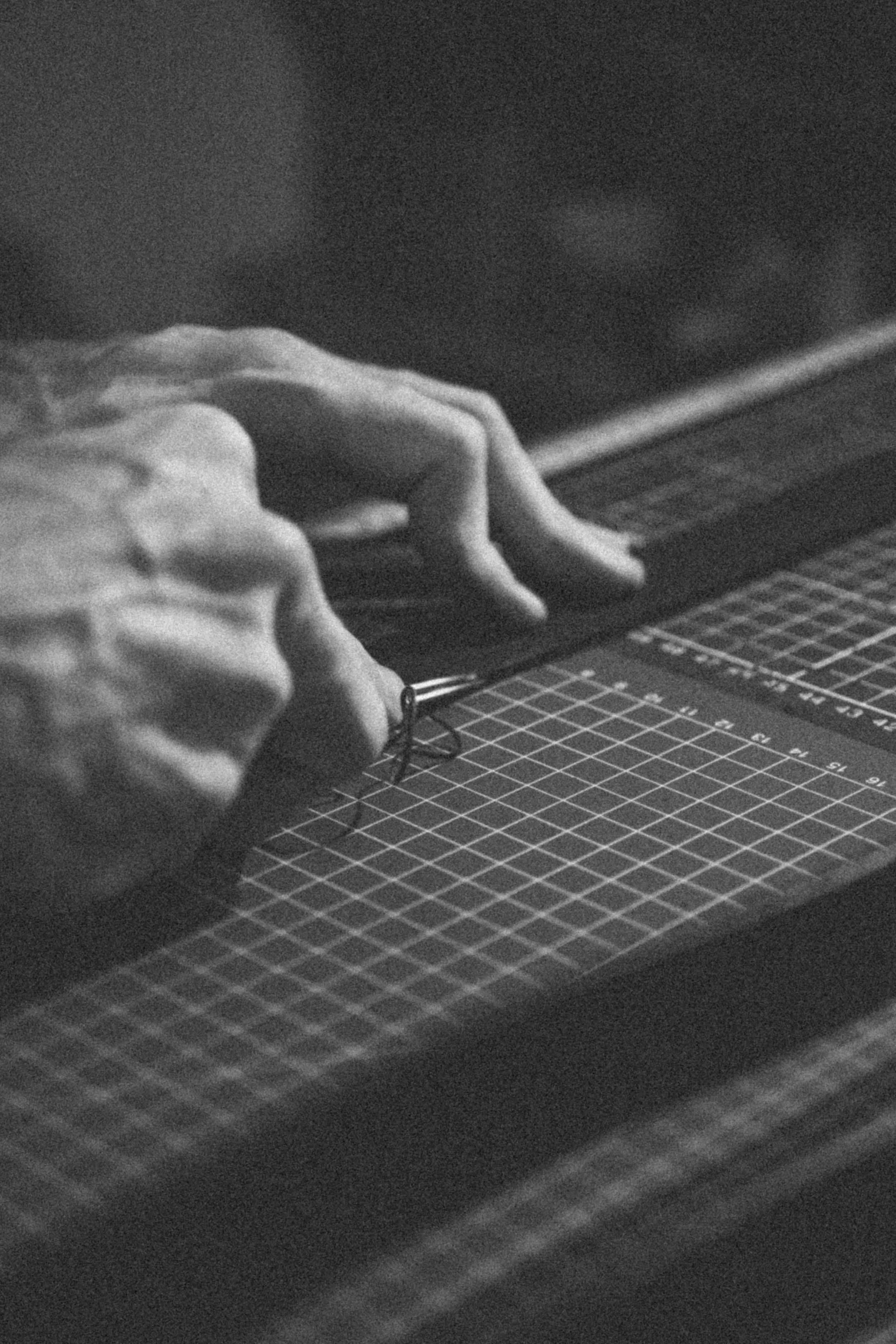 Close-up of person's hand resting on a cutting mat with grid lines, with part of a tool visible near the fingers, in a black and white photo.