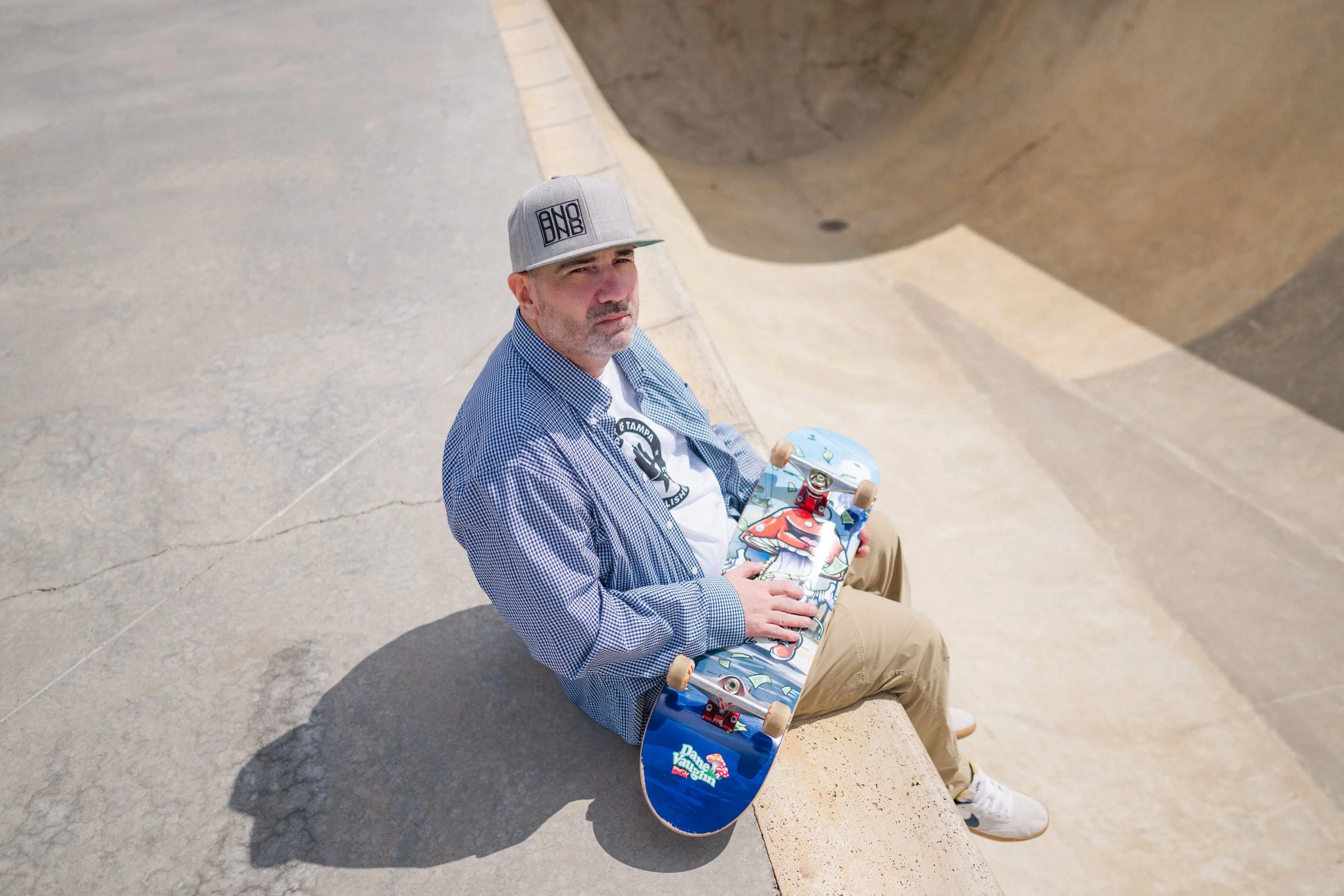 A man sitting on a concrete bench holding a skateboard, wearing a gray baseball cap, a checkered shirt, and beige pants.