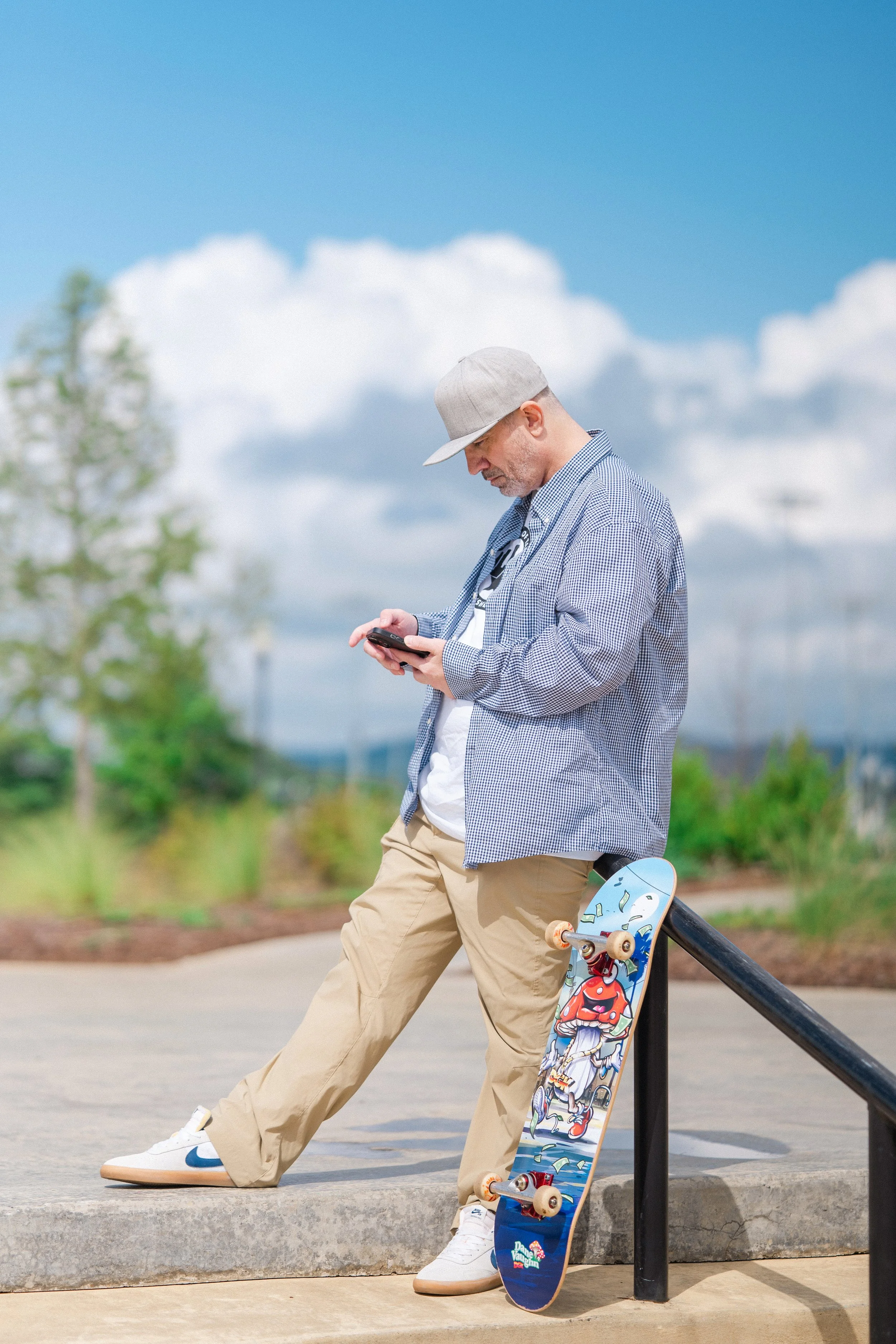 A man wearing a gray cap, checkered shirt, and khaki pants is standing outdoors on a sidewalk, looking at his phone. A skateboard with cartoon graphics leans against a black railing next to him. The background features a blue sky with clouds and some greenery.
