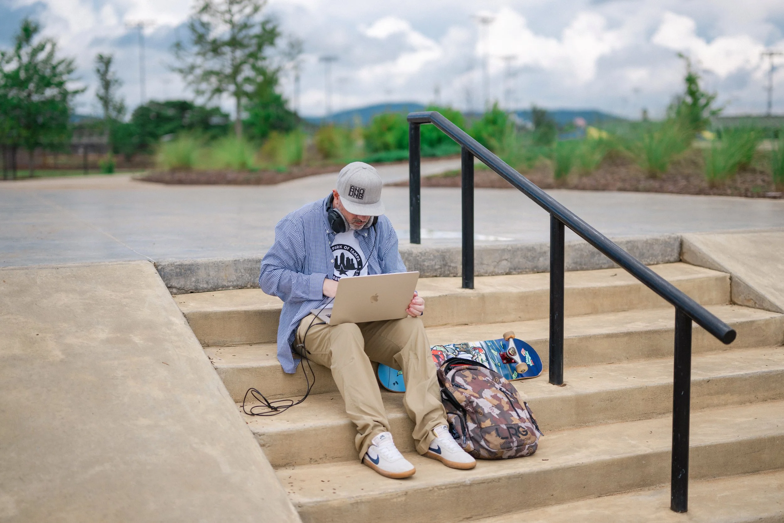 A young person wearing a gray cap with headphones around their neck, a checkered button-up shirt, khaki pants, and white sneakers sitting on concrete stairs with a laptop on their lap, a skateboard and backpack beside them, in an outdoor park setting with trees and cloudy sky in the background.