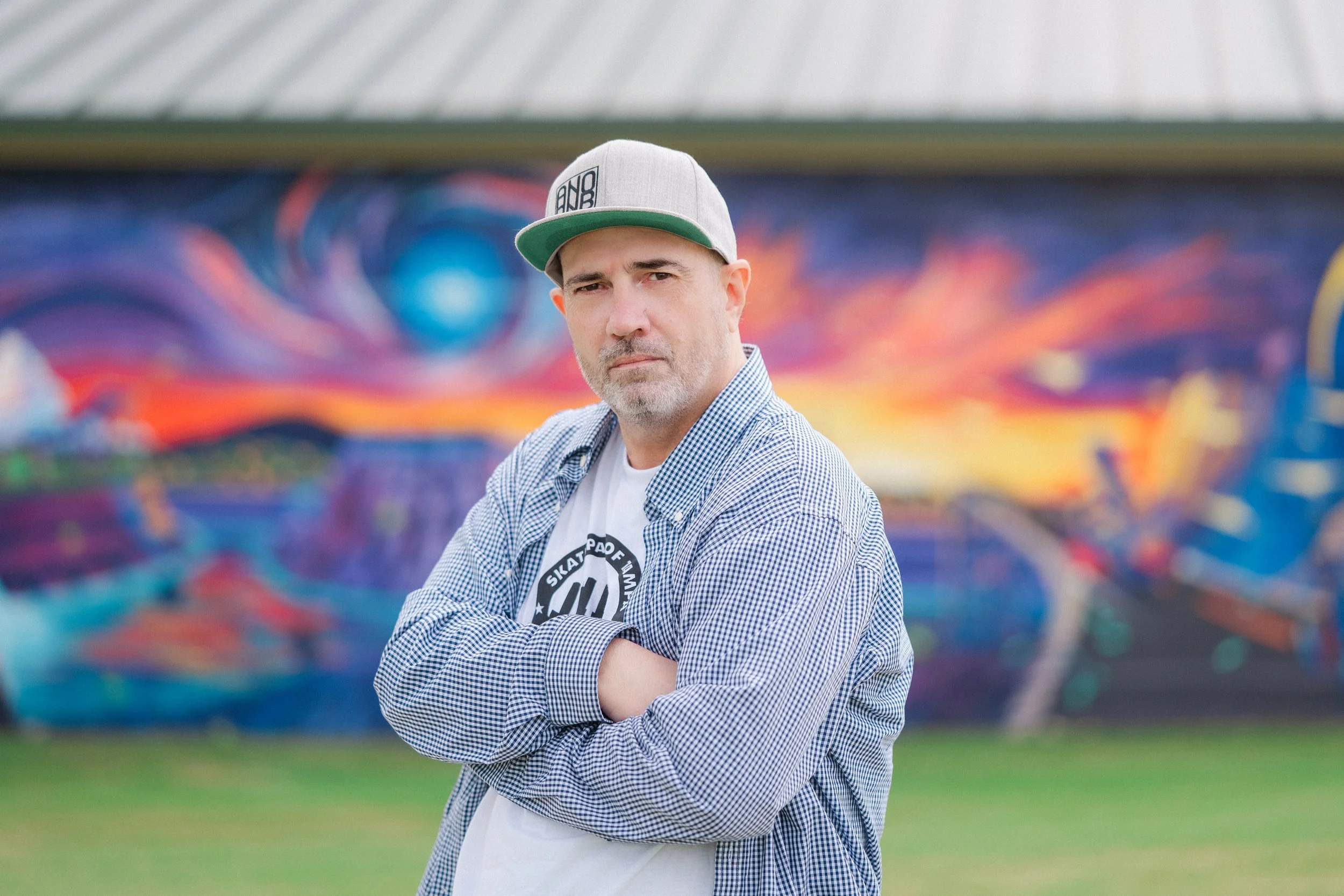 A man with crossed arms wearing a baseball cap and a checkered shirt standing in front of a colorful graffiti mural.