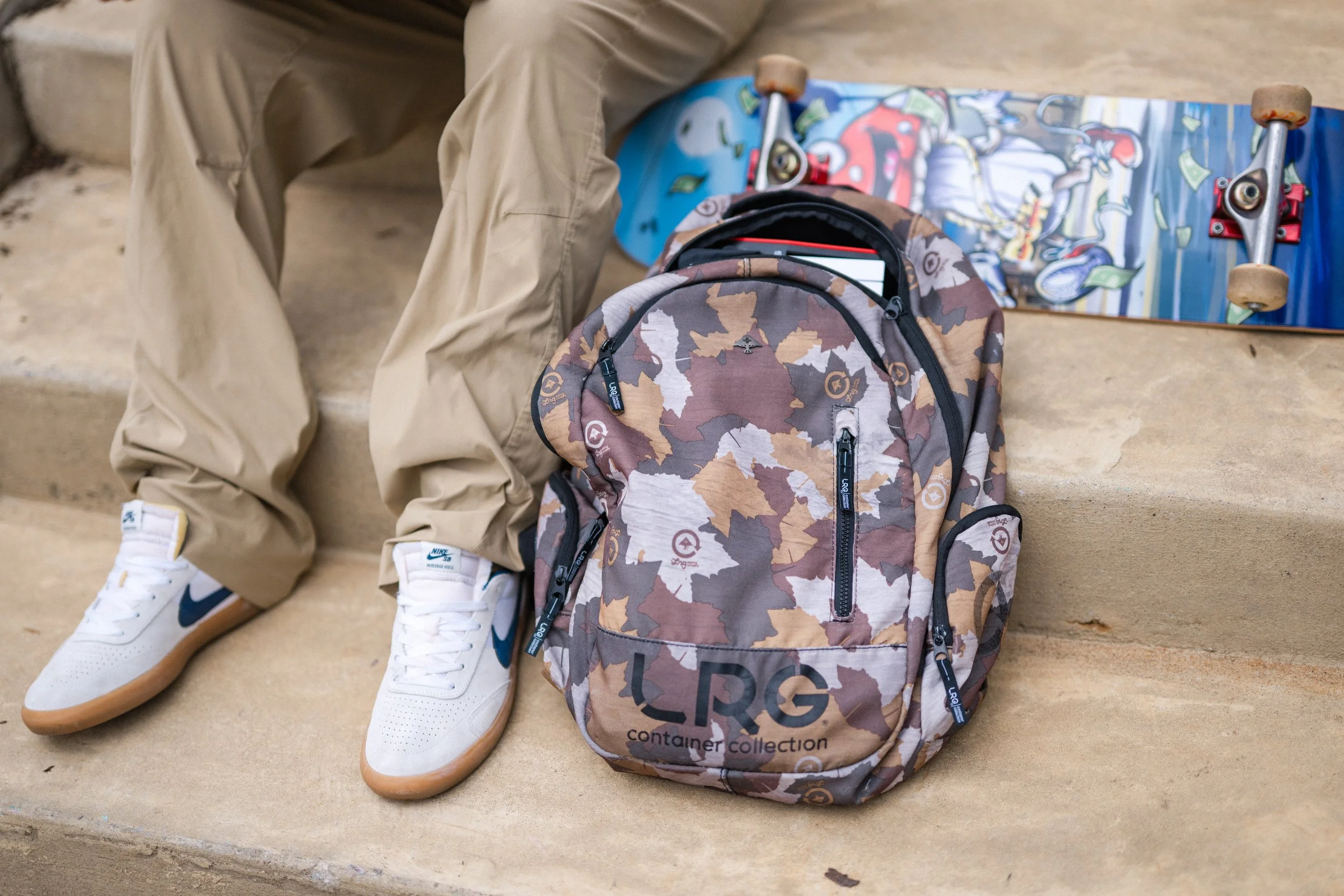 A pair of beige pants, white sneakers, a camouflage backpack, and a skateboard with cartoon graphics are placed on stone stairs.