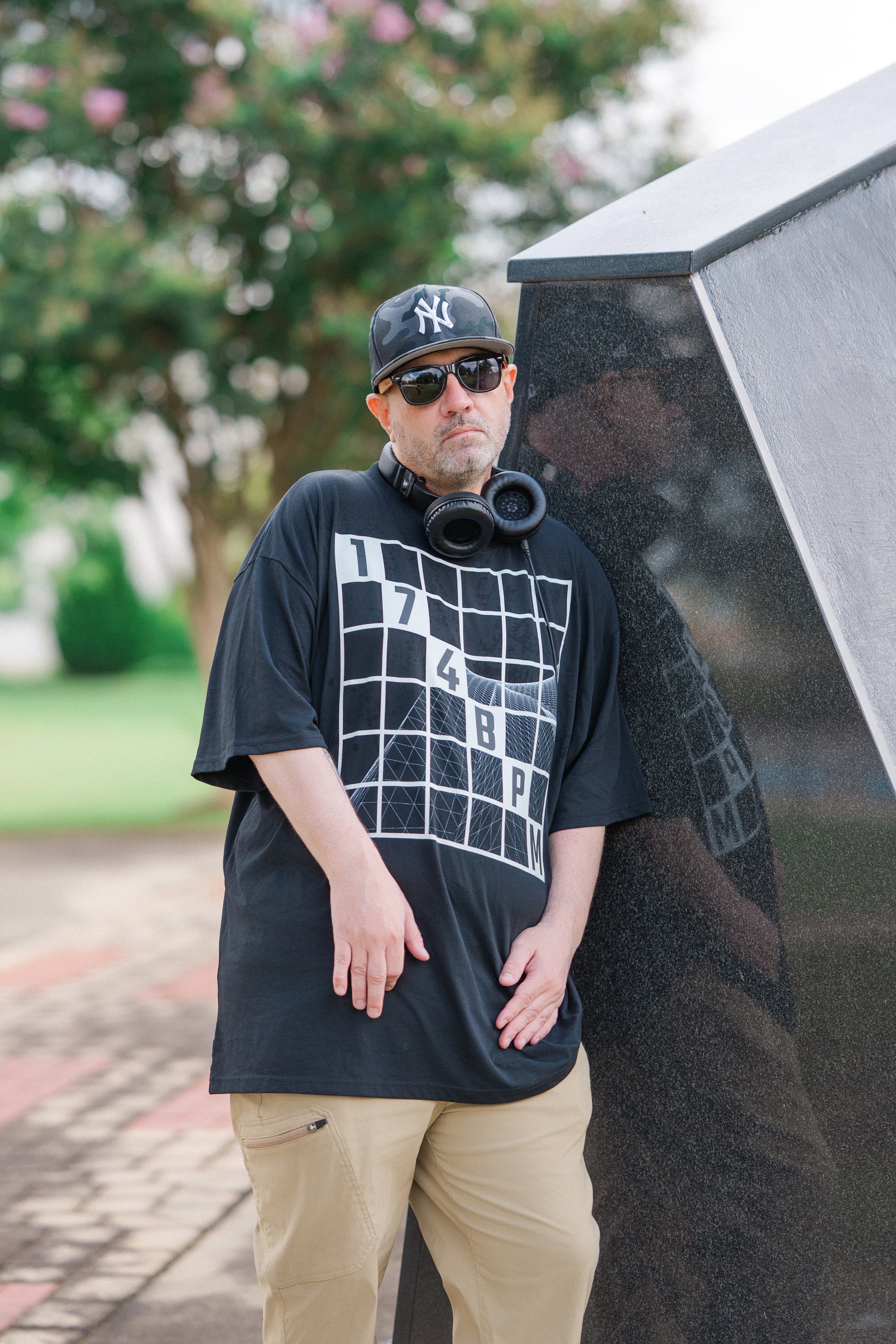A man with a beard, wearing a camouflage New York Yankees cap, black sunglasses, black graphic t-shirt, and beige pants, standing outdoors beside a large black granite monument or sculpture.