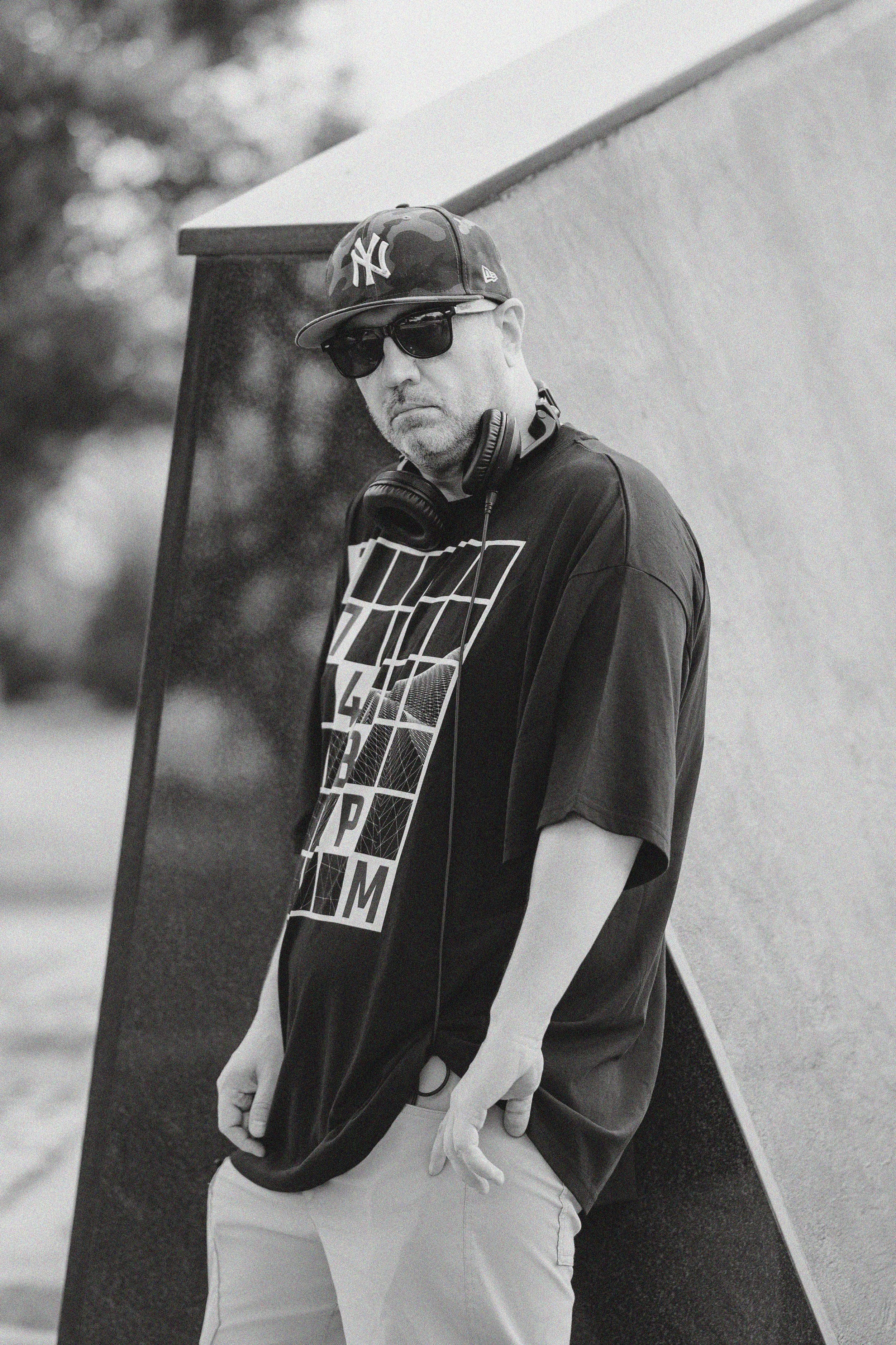 A man wearing sunglasses, a New York Yankees cap, a graphic T-shirt, and headphones around his neck, standing outdoors next to a surfboard.