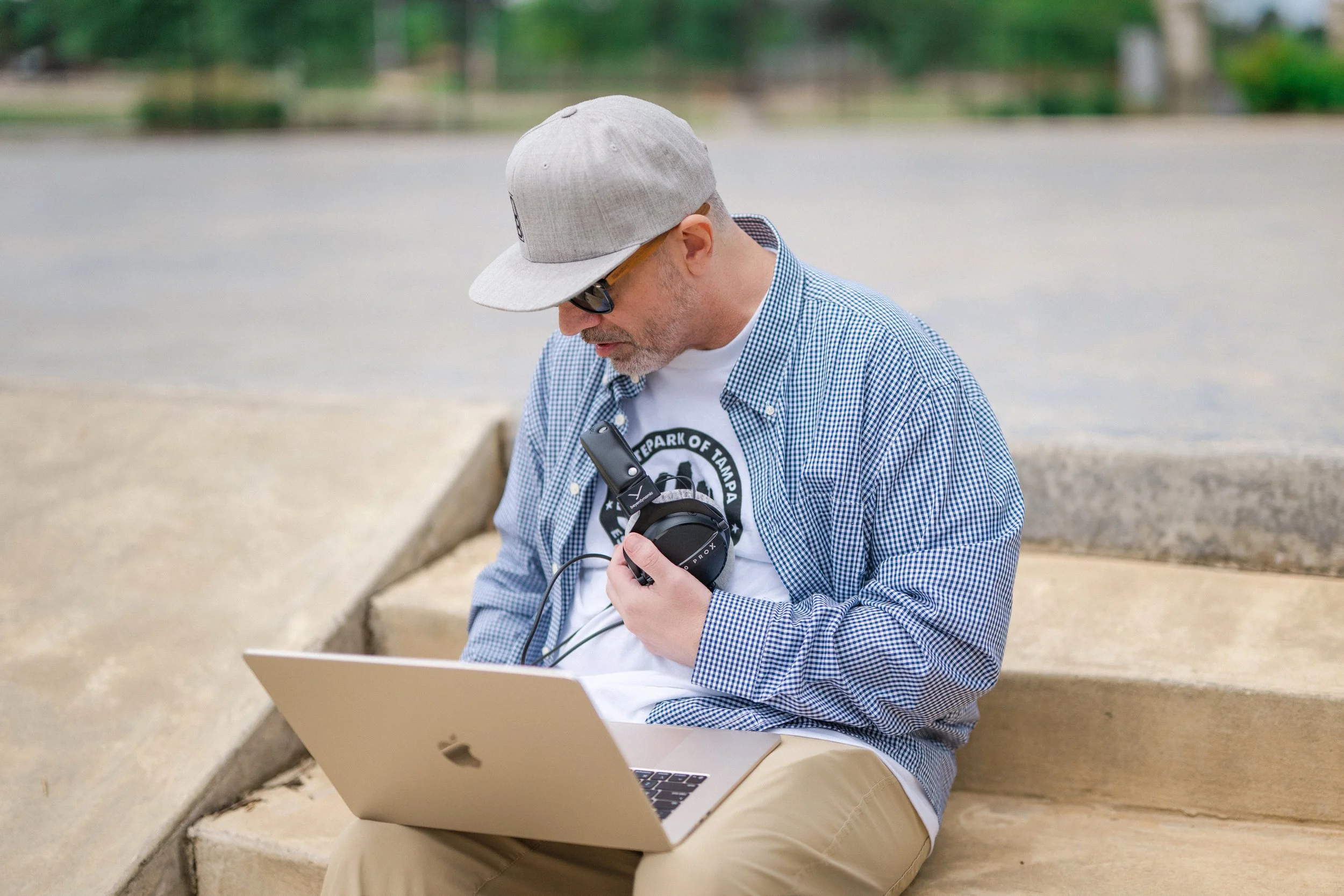 A man sitting outdoors at a bench, wearing a gray cap, sunglasses, a checkered shirt, and beige pants. He is looking at his phone and has a laptop open in front of him.